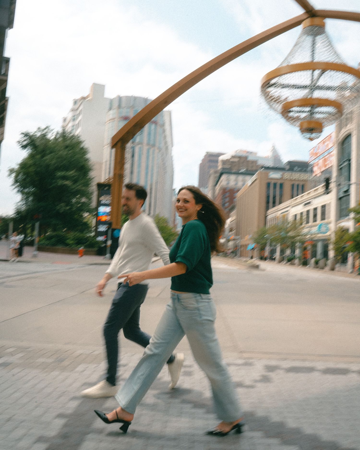 A smiling couple walks hand-in-hand on a city street, with urban buildings and a decorative arch in the background.