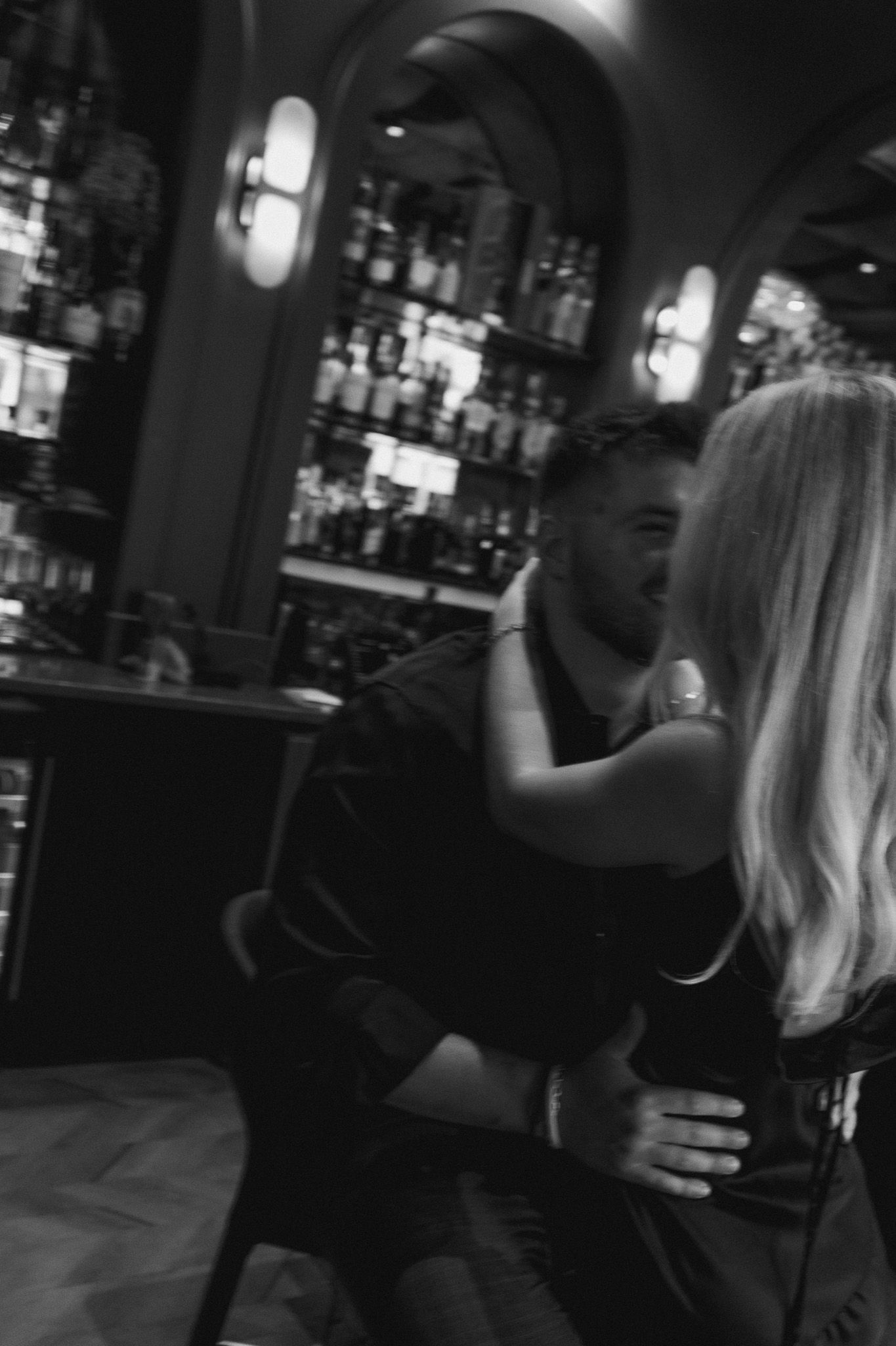 Black and white photo of a couple embracing in a dimly lit bar with liquor bottles on shelves in background.