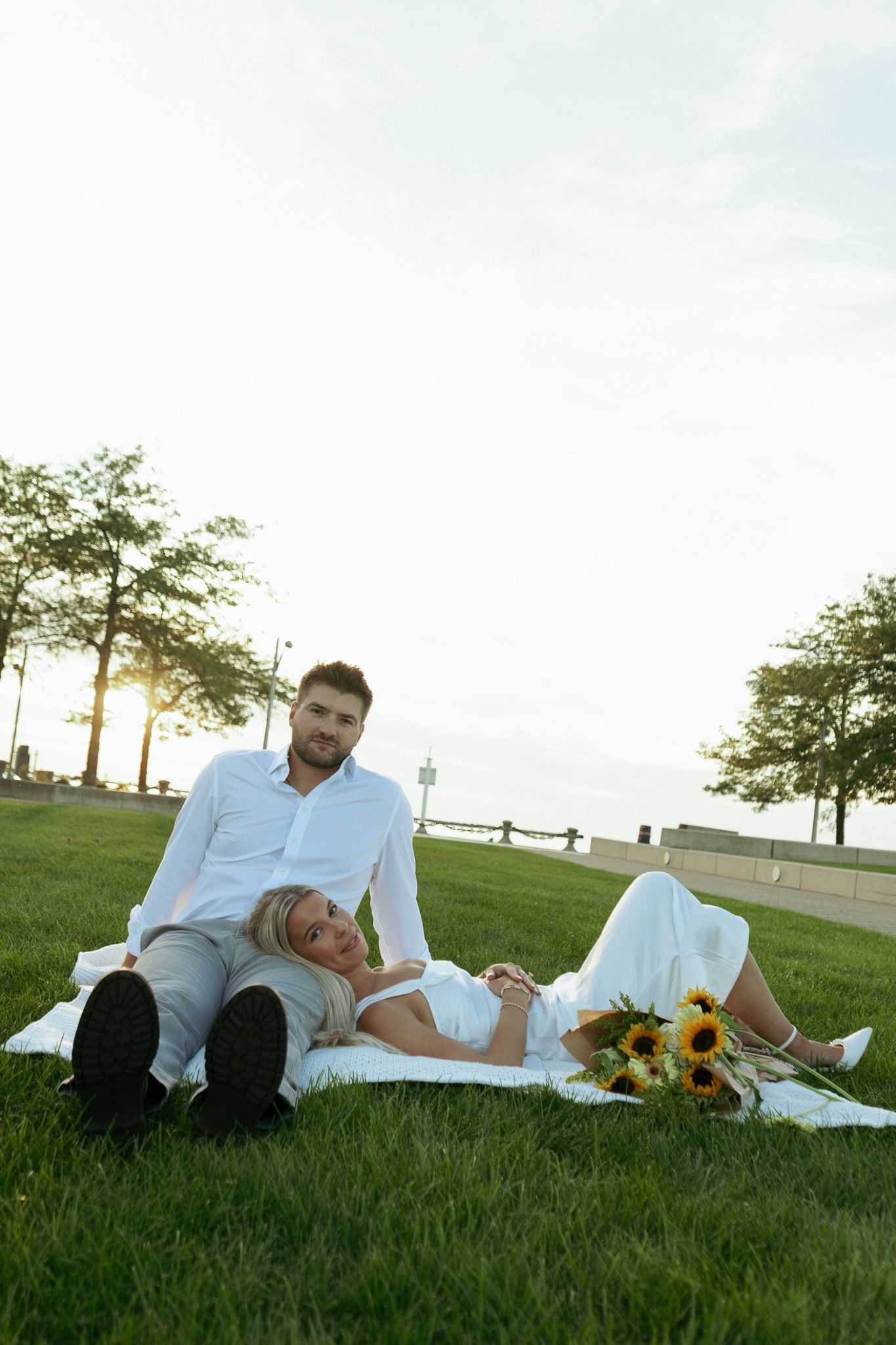 Bride and groom in white attire relaxing on green grass with sunflowers during golden hour outdoor wedding photo.