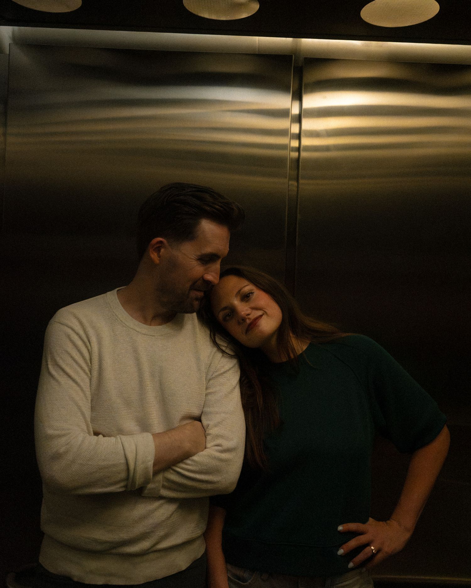 A couple shares a tender moment leaning together in a dimly lit elevator, woman in green top resting head on mans shoulder.