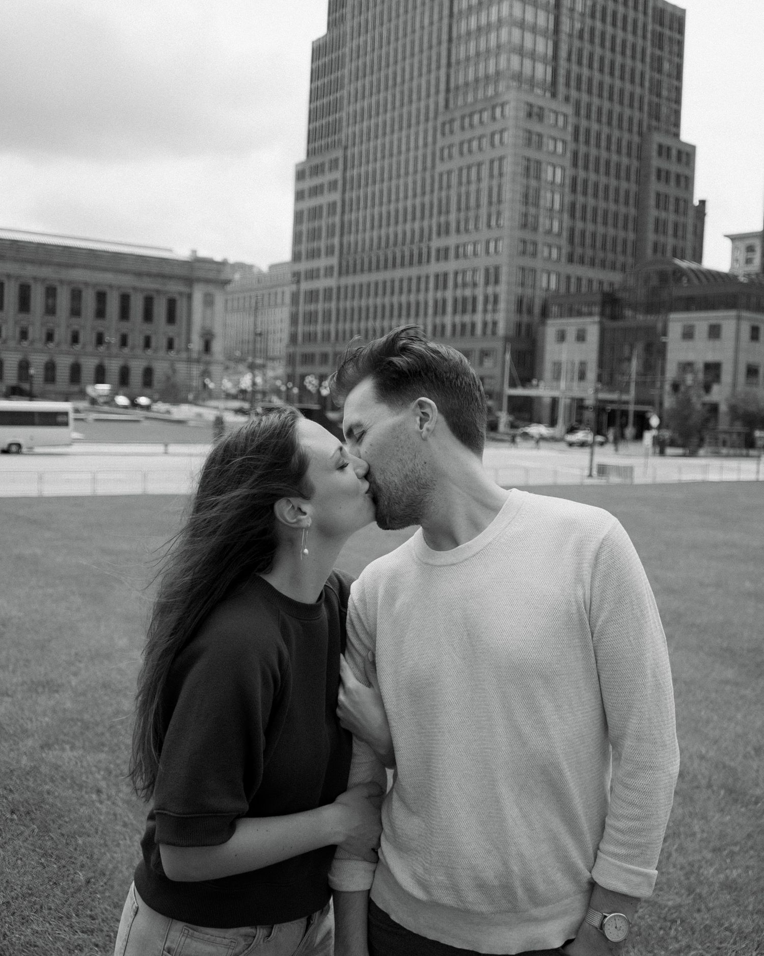 Black and white photo of a couple sharing a romantic kiss outdoors with a city skyline in the background.