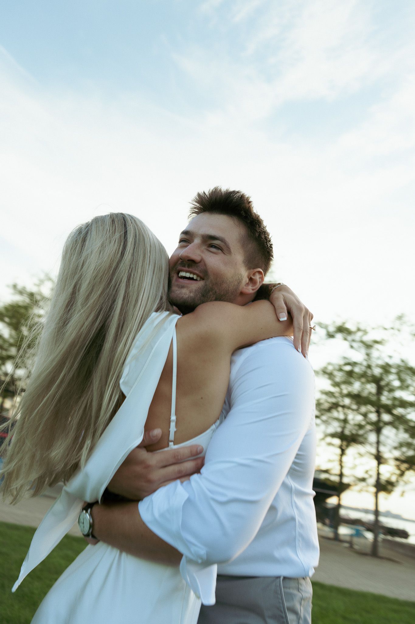 Happy couple embracing outdoors, smiling man in white shirt hugging blonde woman in white dress against blue sky.