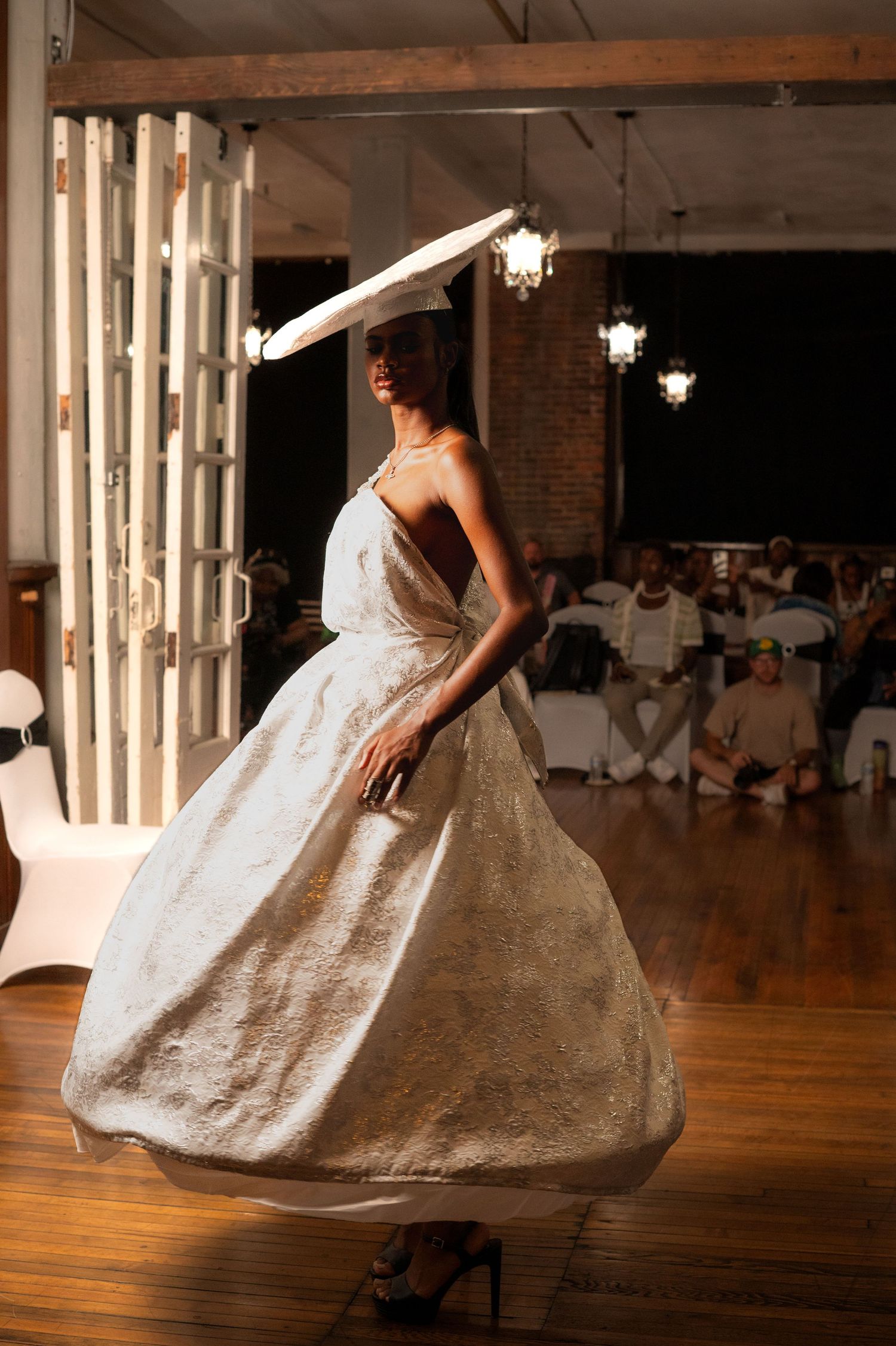 Model walks runway in gold ballgown with wide-brim hat at indoor fashion show.