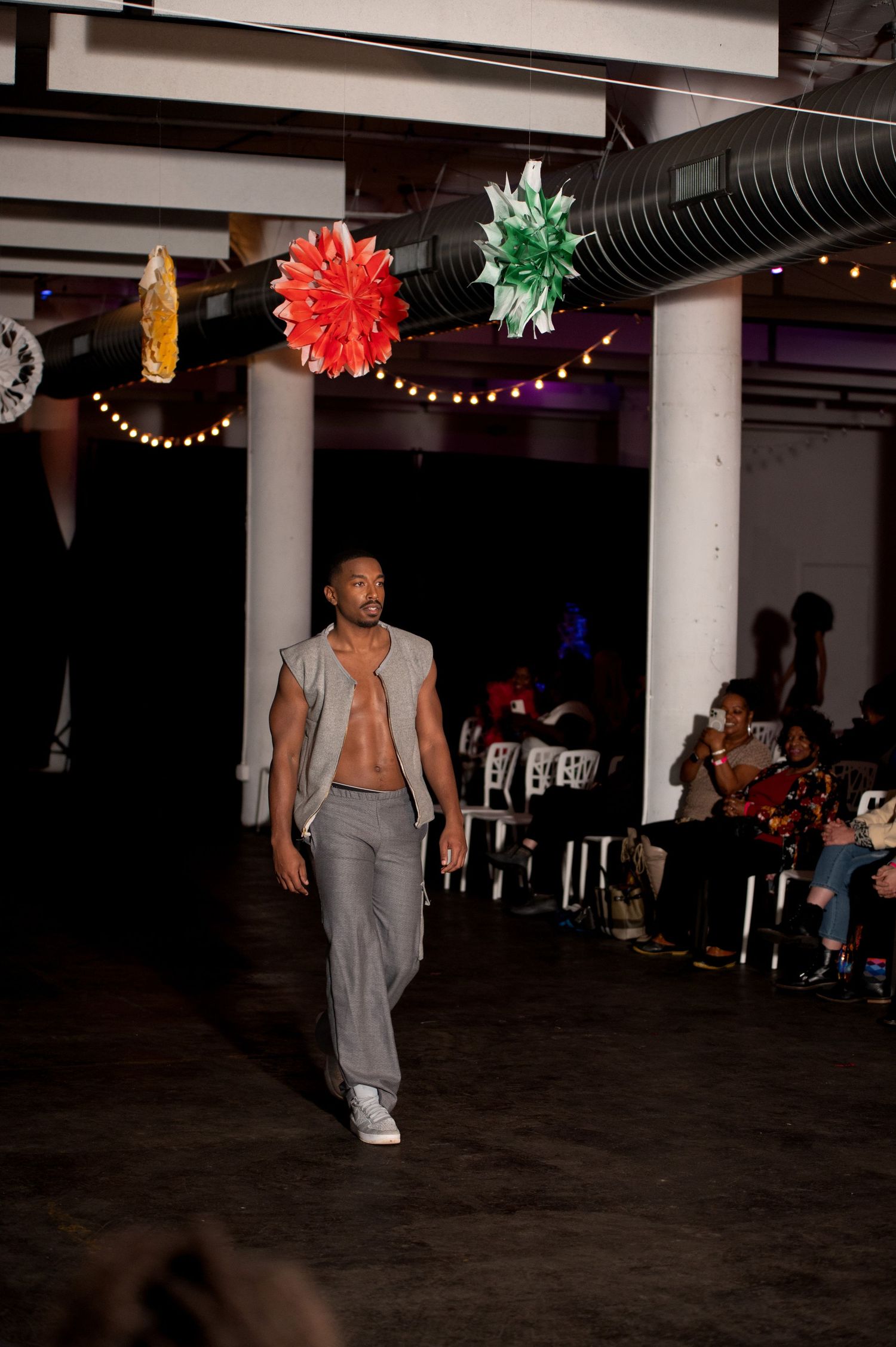 Male model walks runway in open gray vest and pants at indoor fashion show.
