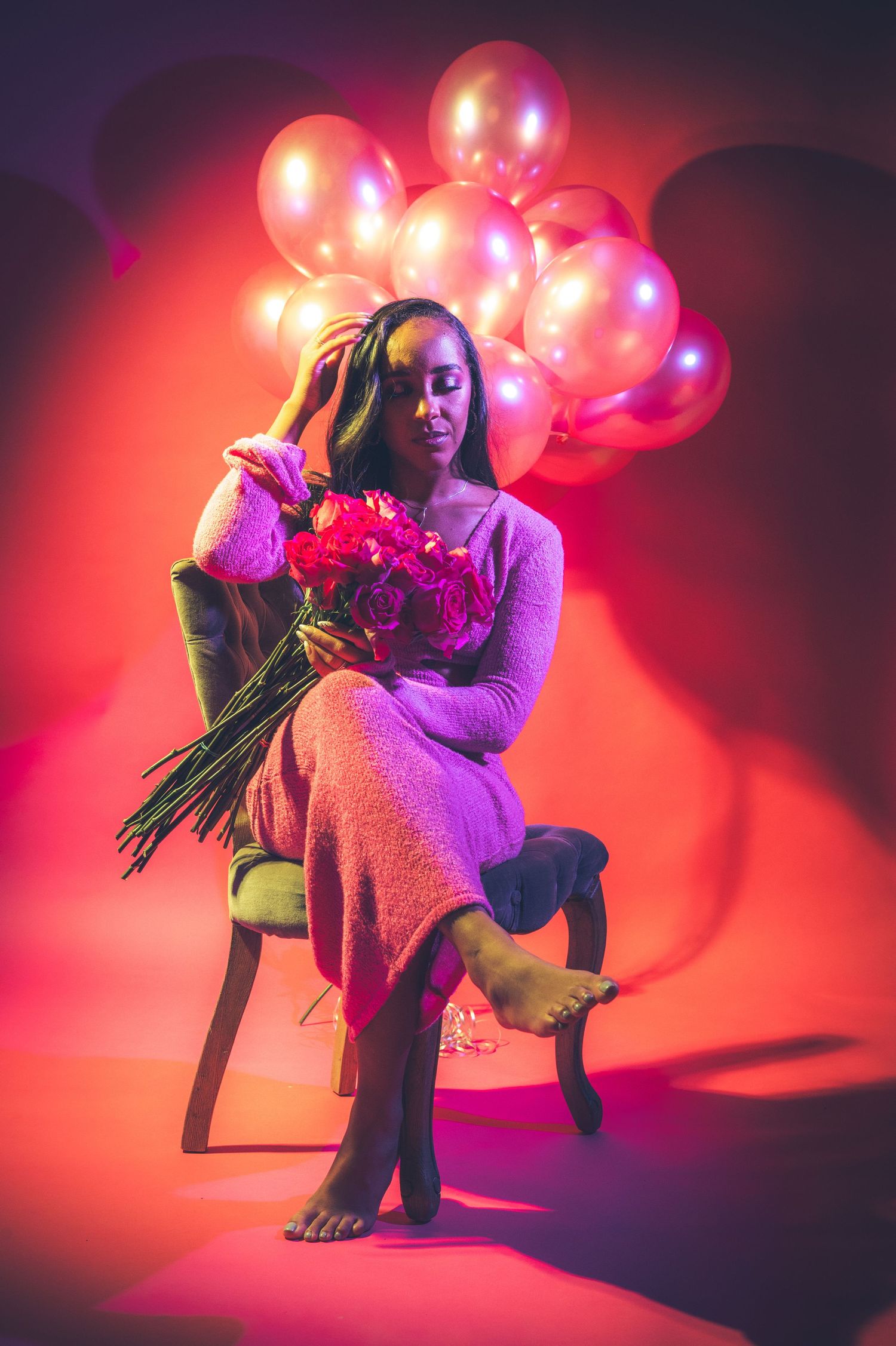 Woman in pink dress sits barefoot holding roses with illuminated pink balloons behind her.