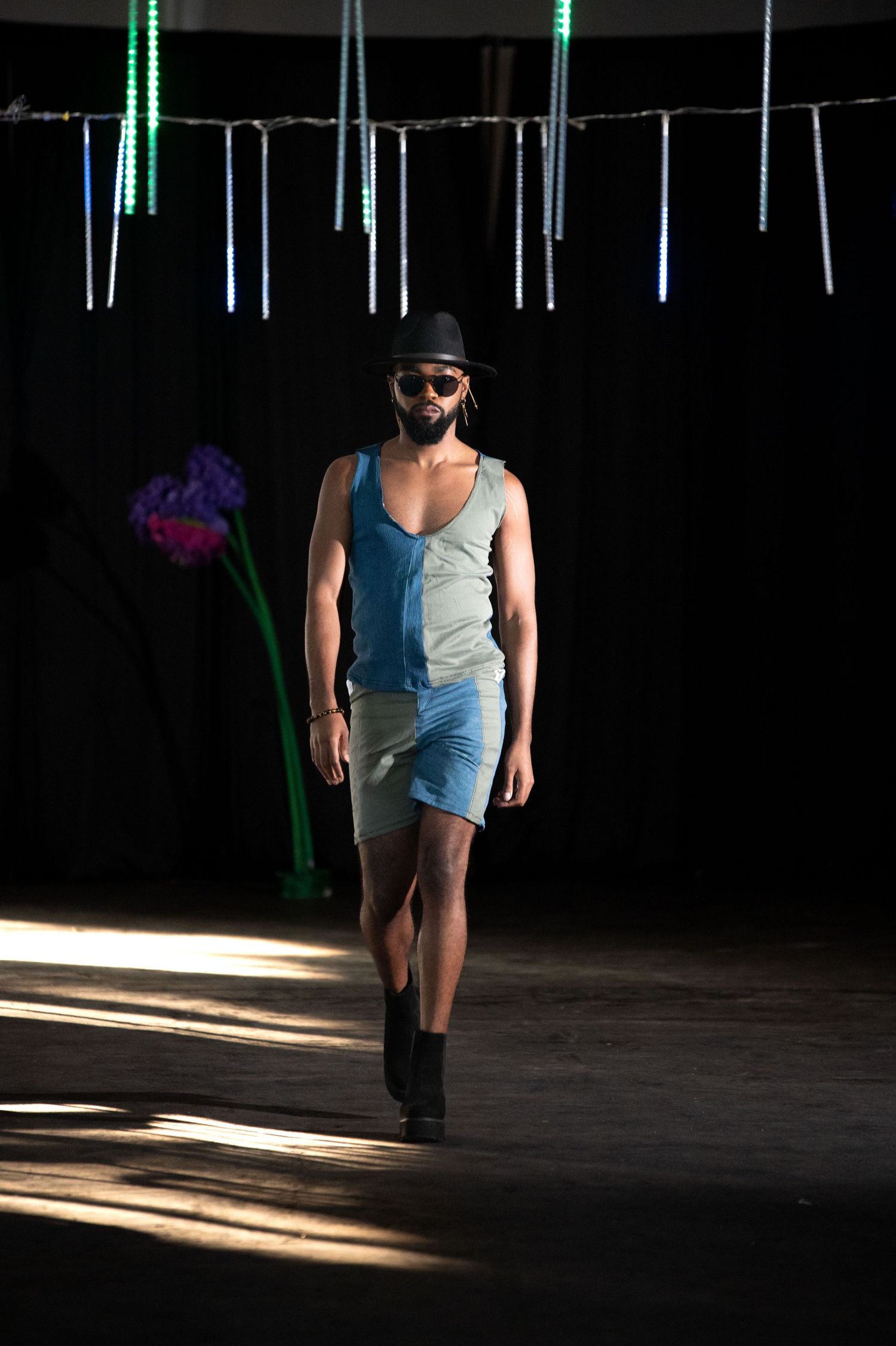 Male model walks runway in denim shorts and tank top with black hat and sunglasses at fashion show.