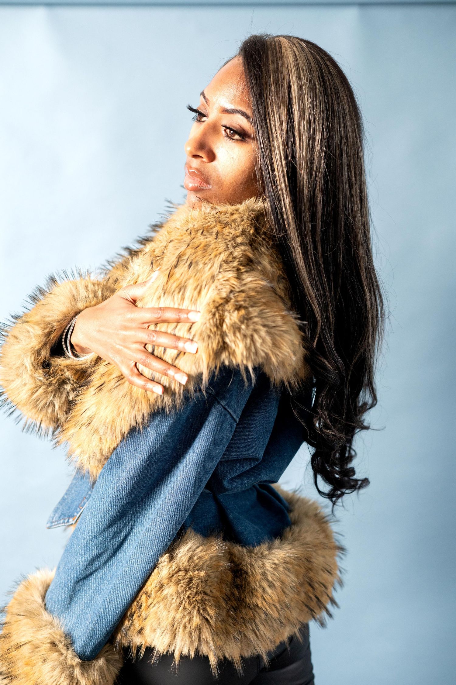 Woman in fur and denim jacket poses elegantly against blue background.