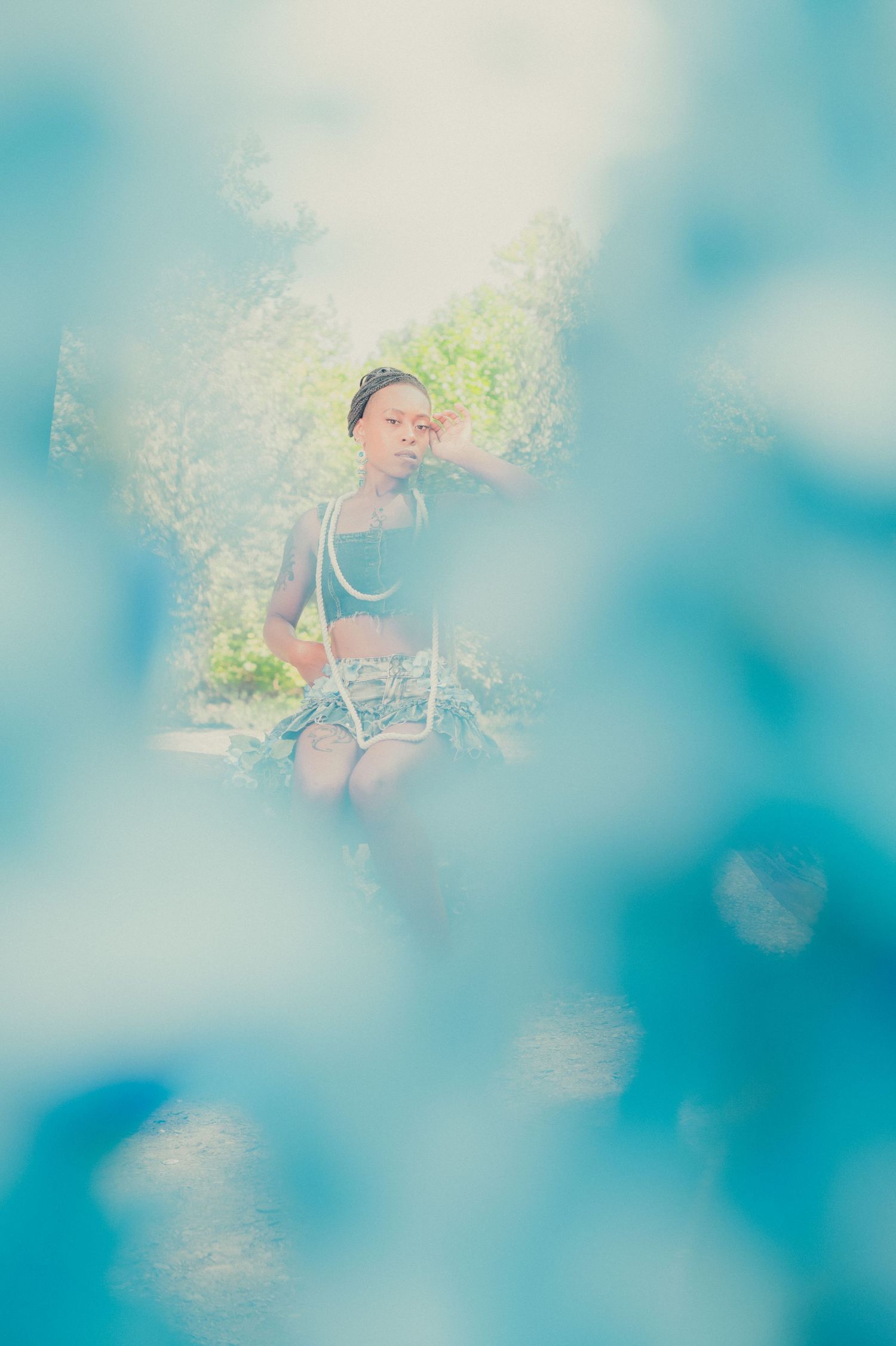 Young woman in blue crop top sits outdoors framed by soft blurred blue bokeh foreground.
