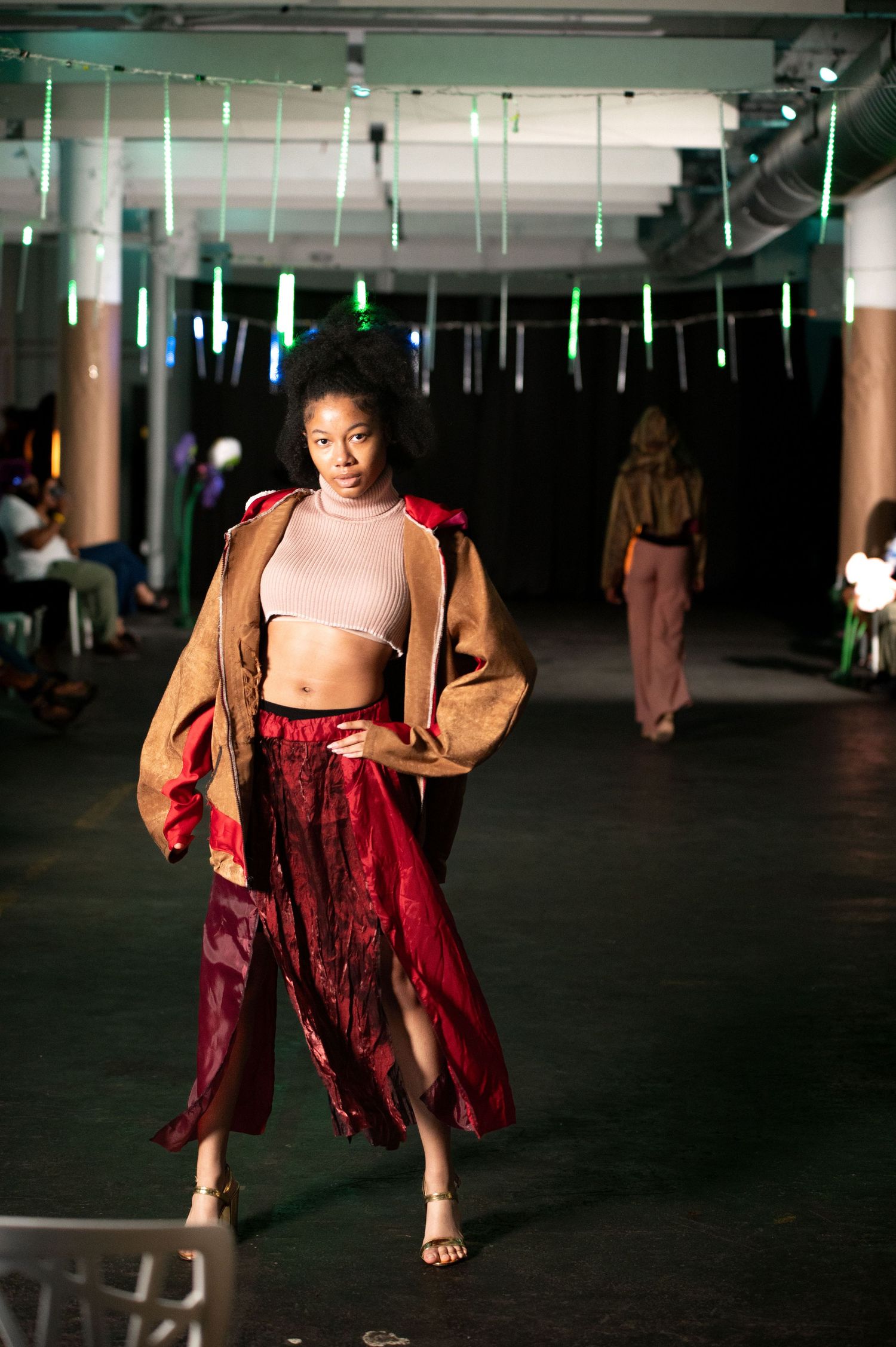 Female model wearing burgundy skirt and tan jacket walks fashion show runway with confidence.