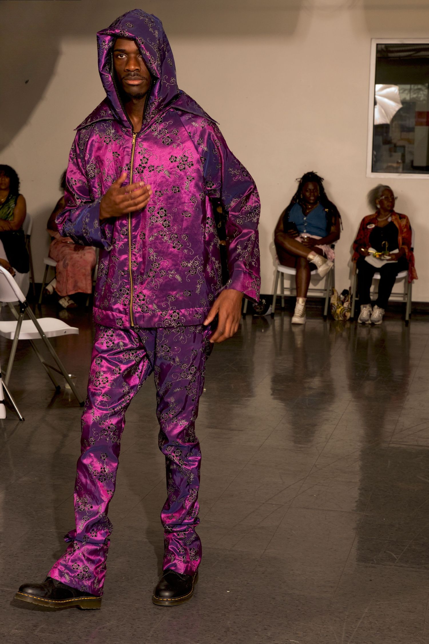 Male model wearing purple and pink floral hooded tracksuit walks fashion show runway.
