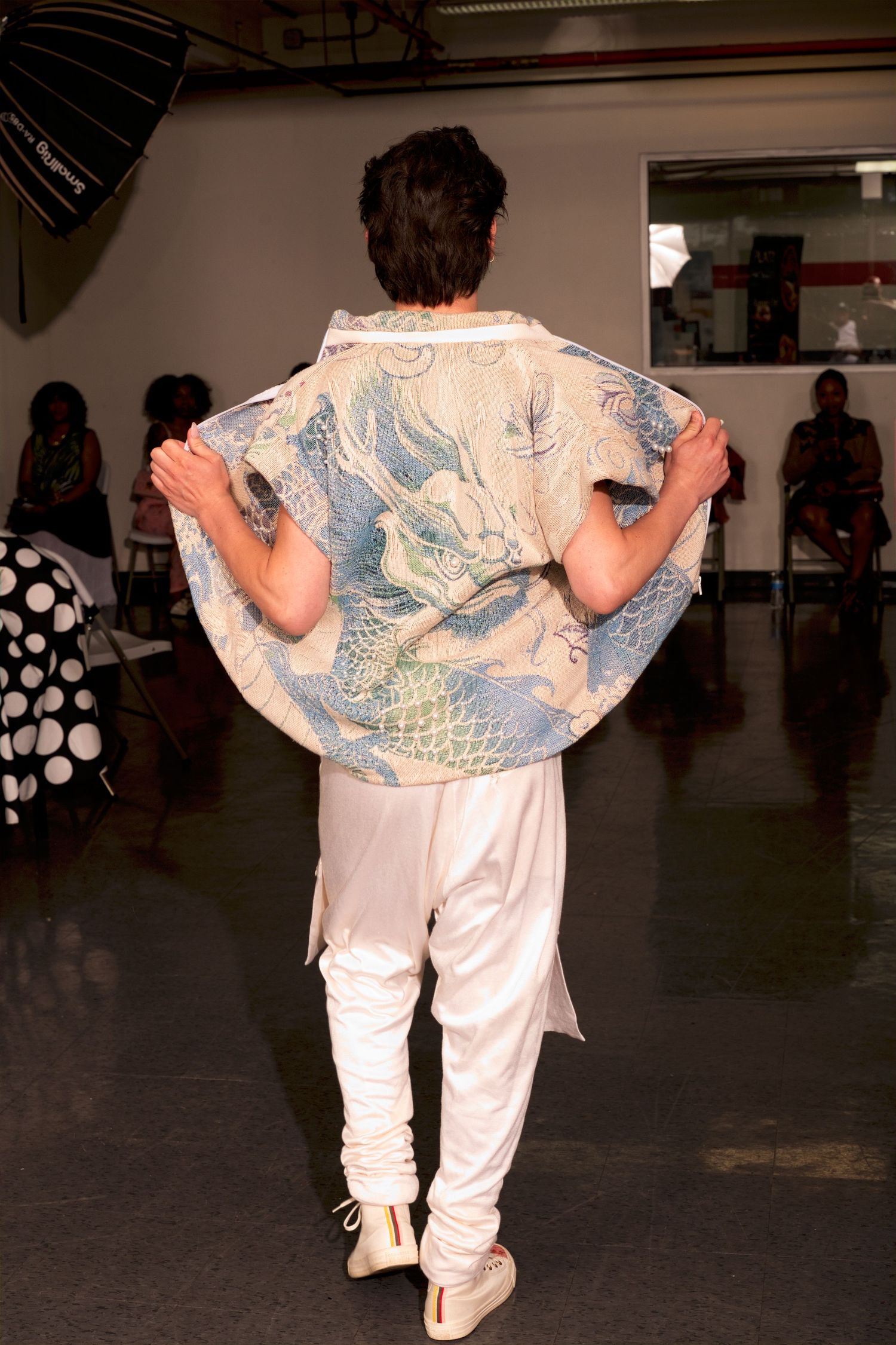 Model displays dragon-printed kimono jacket with white pants from behind on fashion runway.