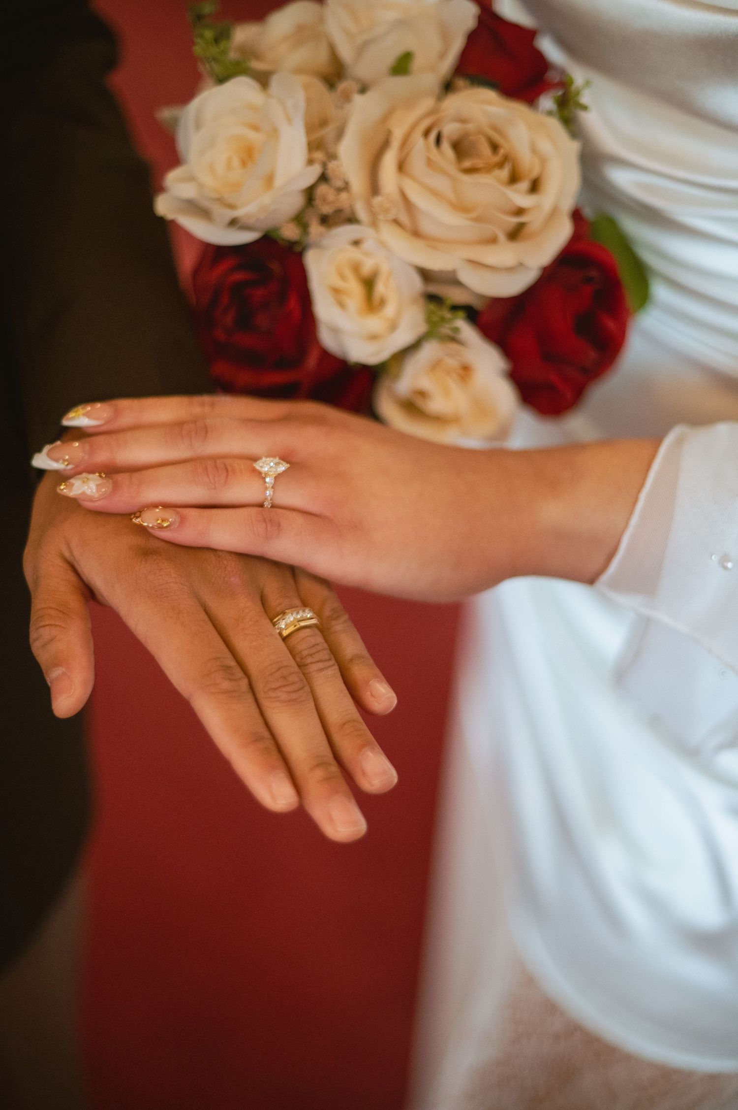 Bride and groom hands showing wedding rings with a bouquet of red and white roses in the background.