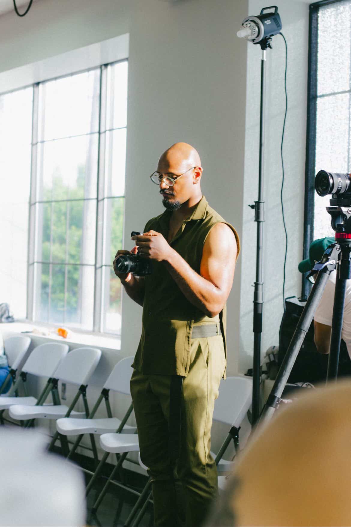 Bald photographer in olive outfit holding camera in bright professional photo studio.