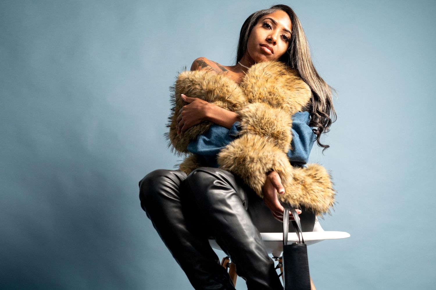 Woman in denim jacket with fur trim sits on stool posing confidently.