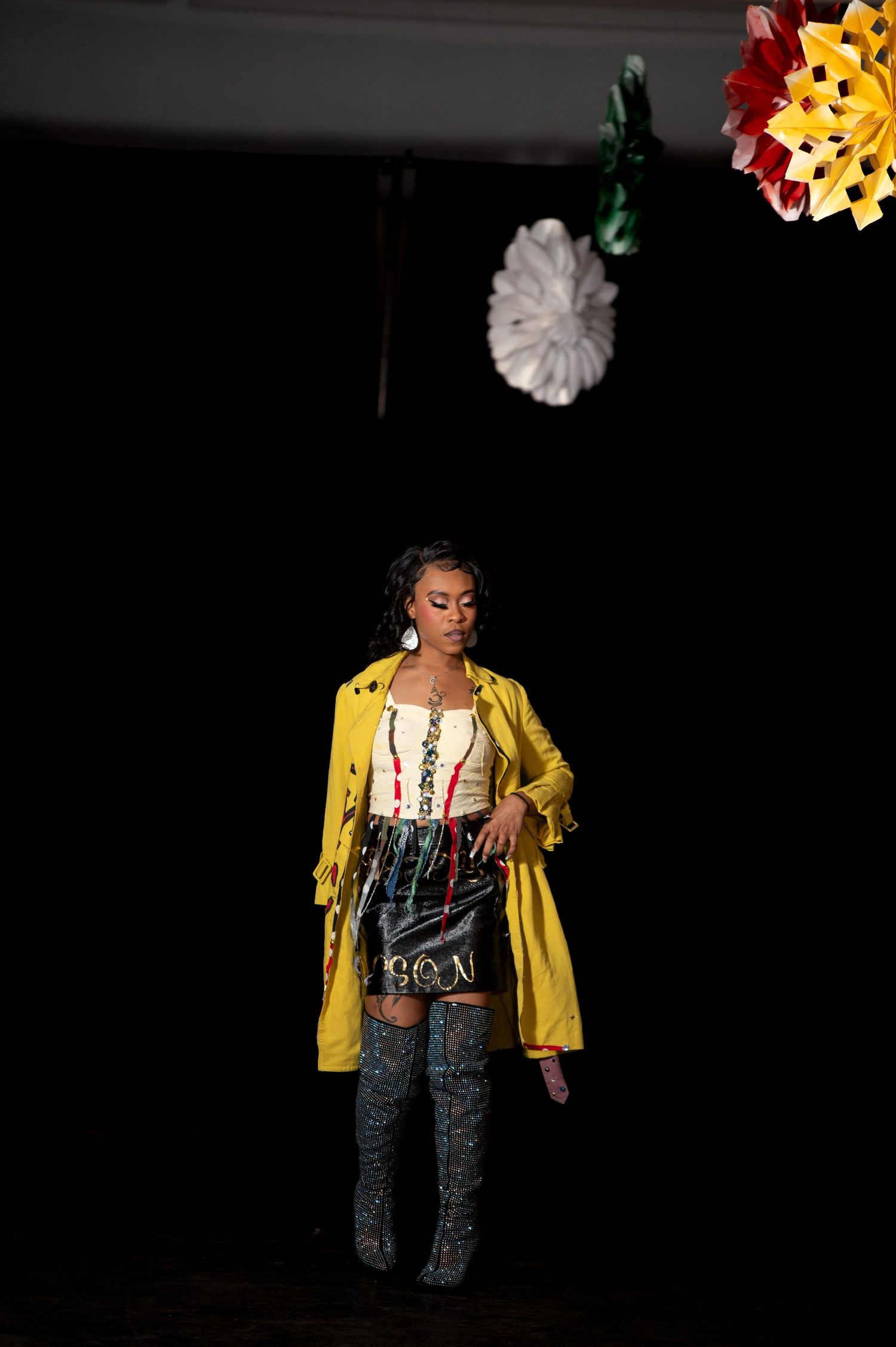 Model poses in yellow trench coat, corset top, and thigh-high boots at fashion show.
