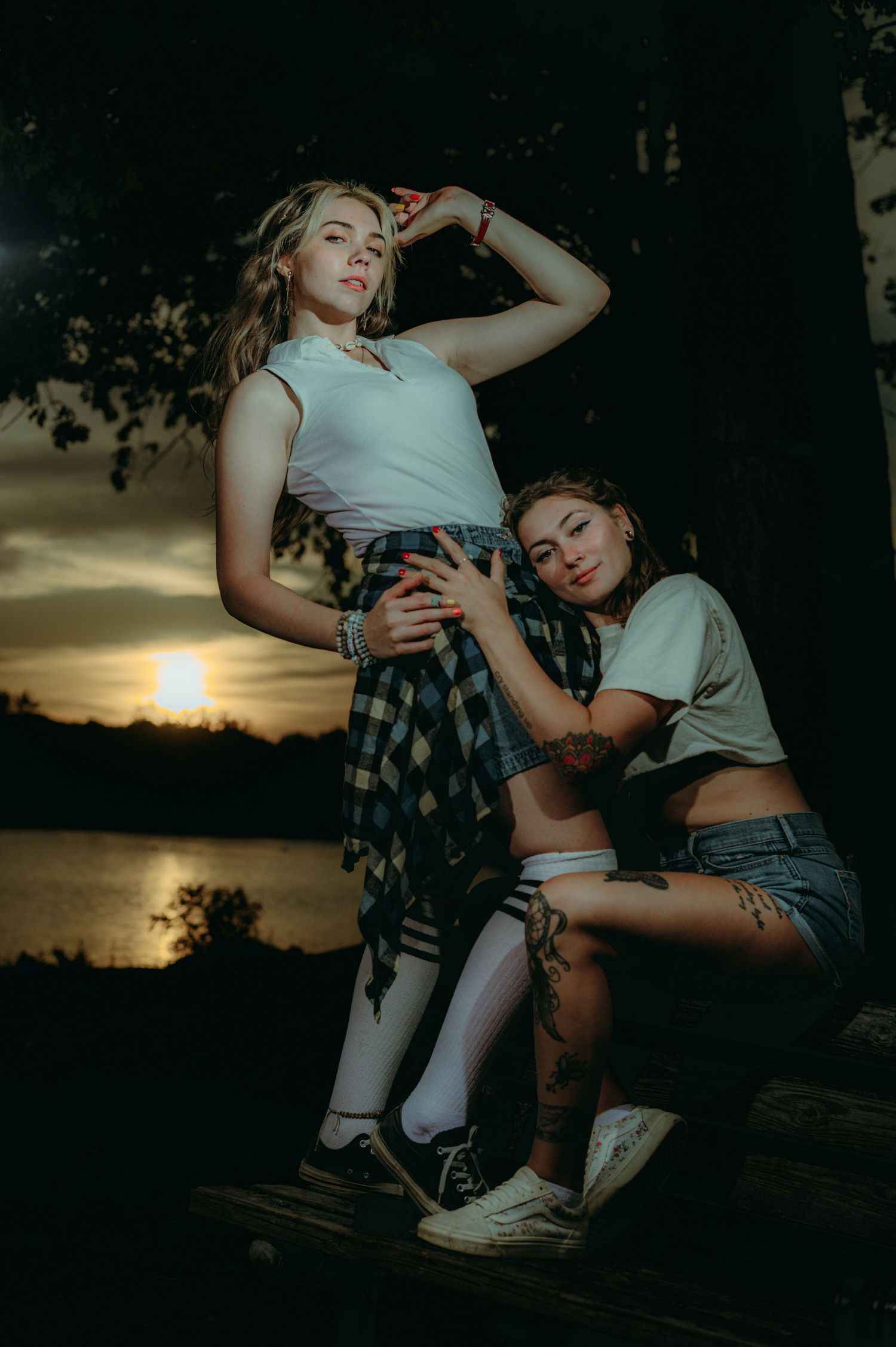 Two women pose dramatically at sunset near a lake, wearing casual white tops and plaid skirts.