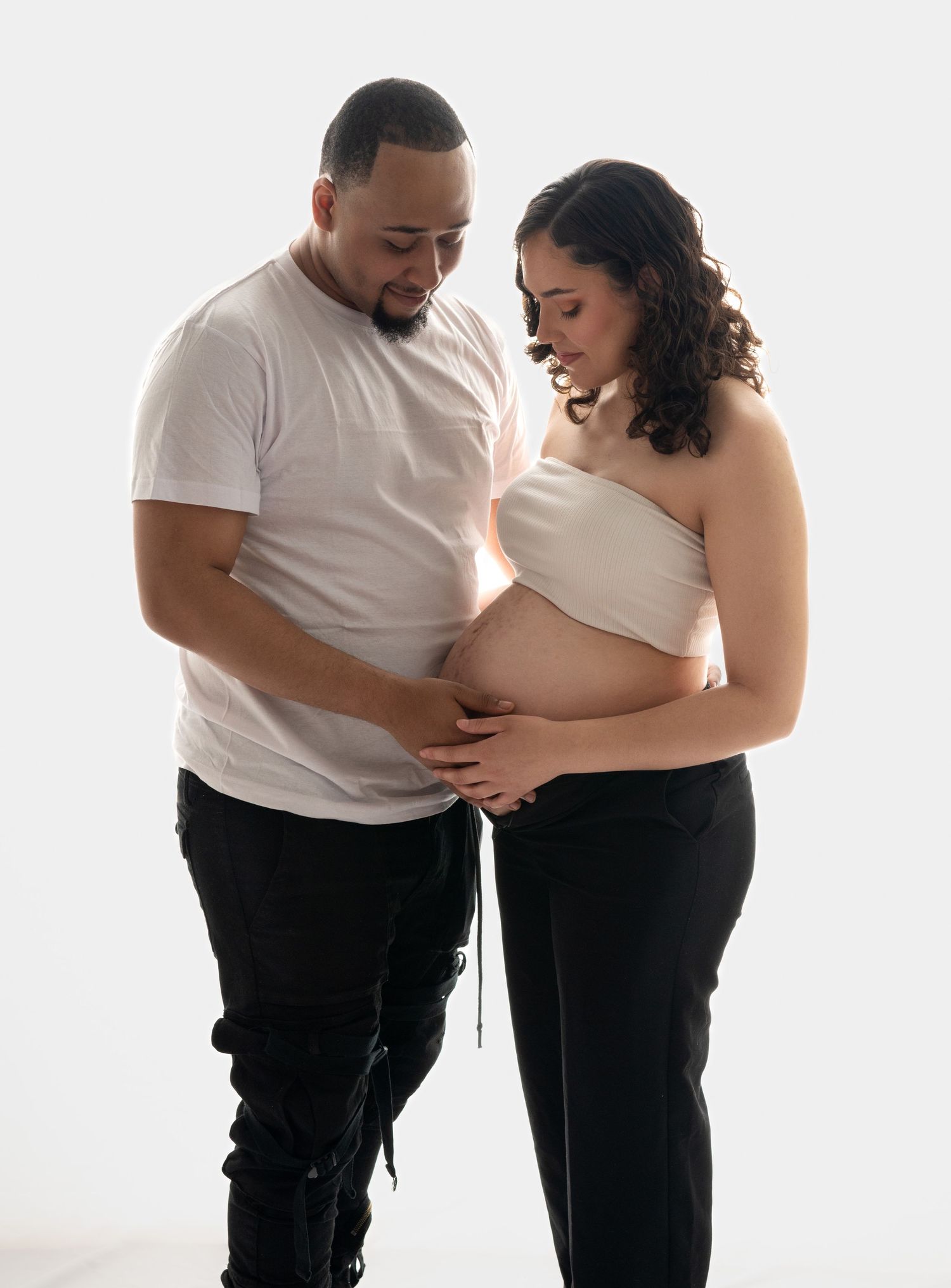 Expecting couple tenderly cradles pregnant belly together in a clean white studio maternity portrait.