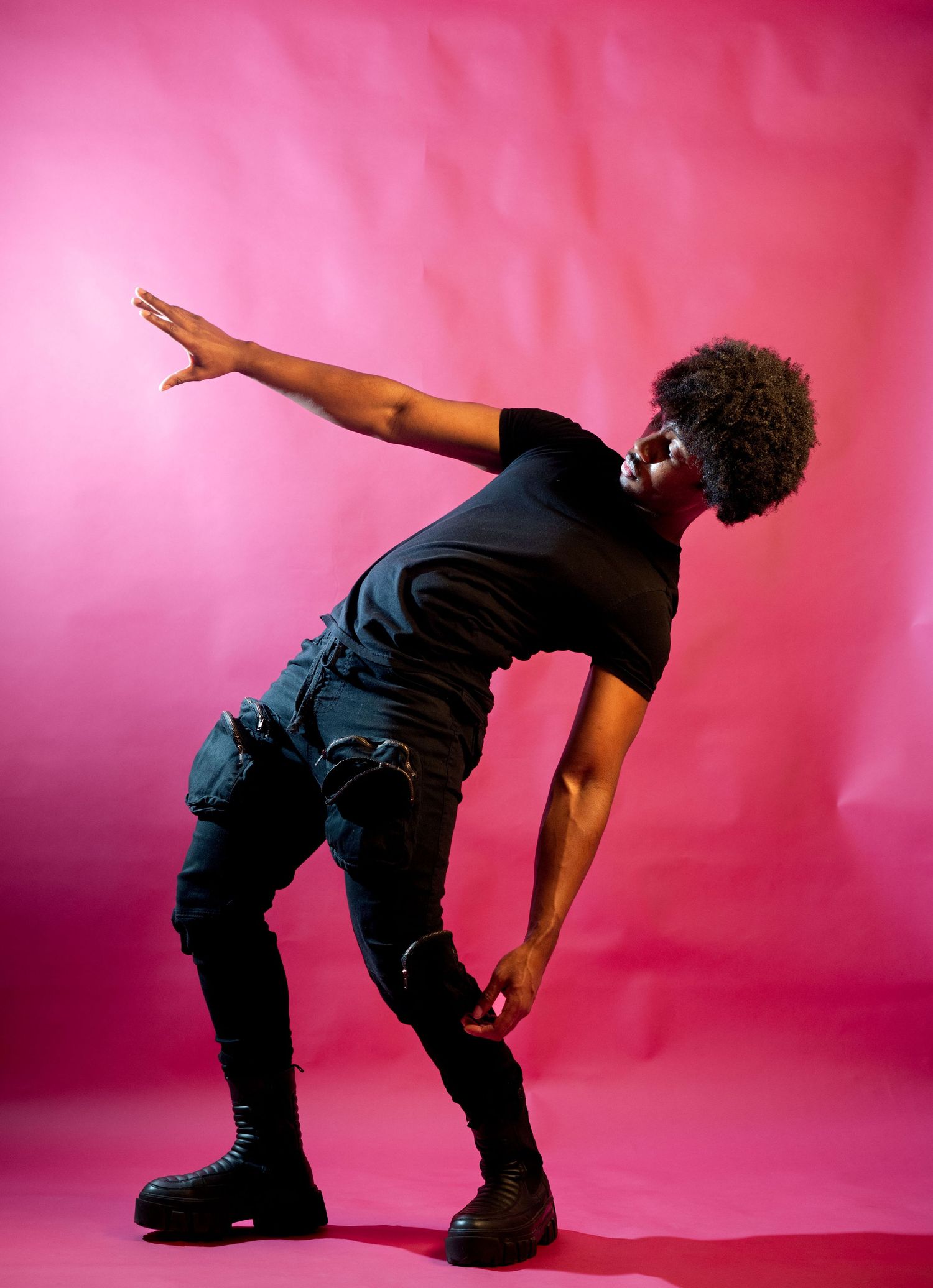 Man with afro in all-black outfit strikes dynamic pose on pink backdrop.