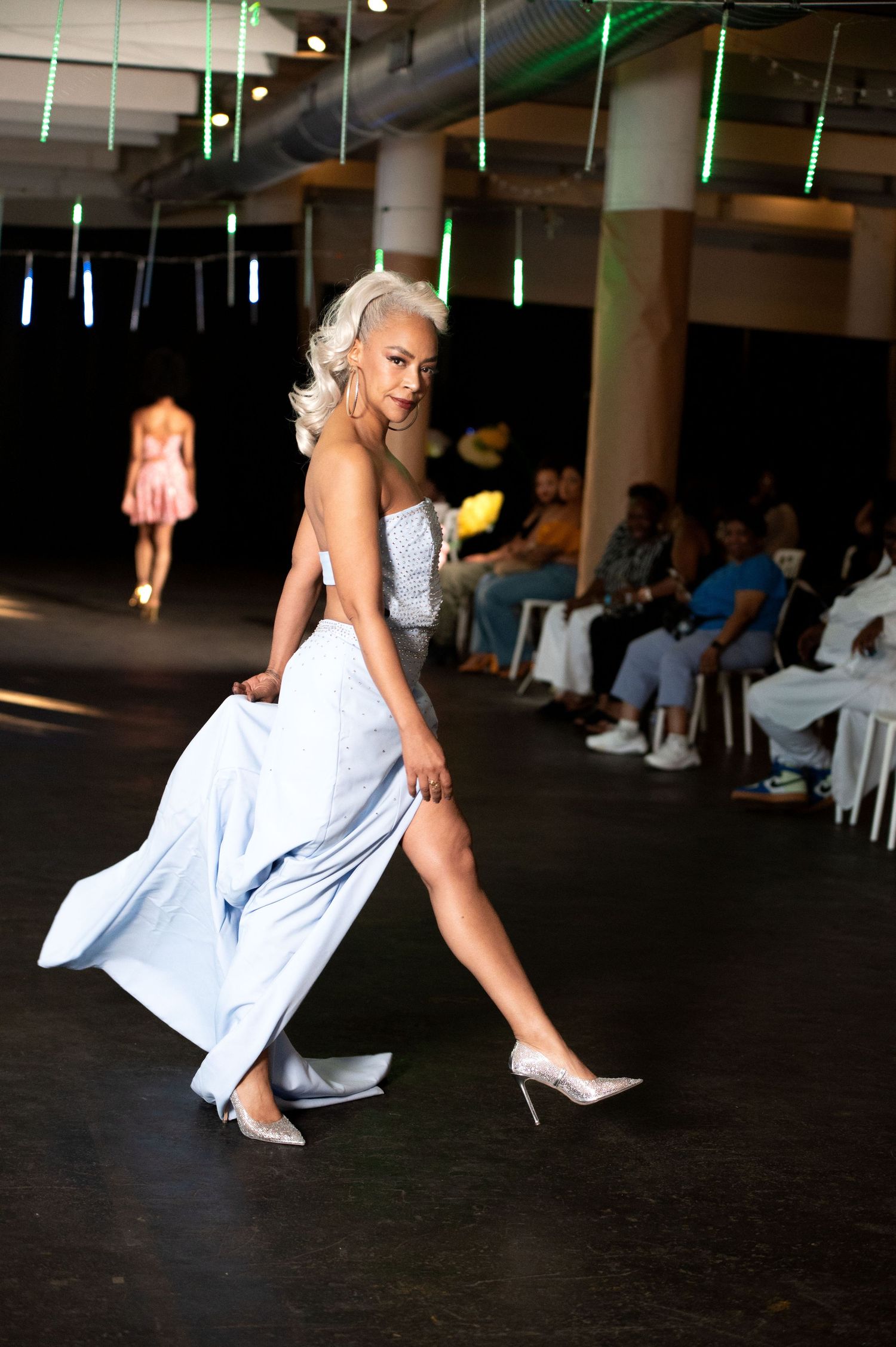 Female model in flowing light blue gown and silver heels struts down fashion show runway.
