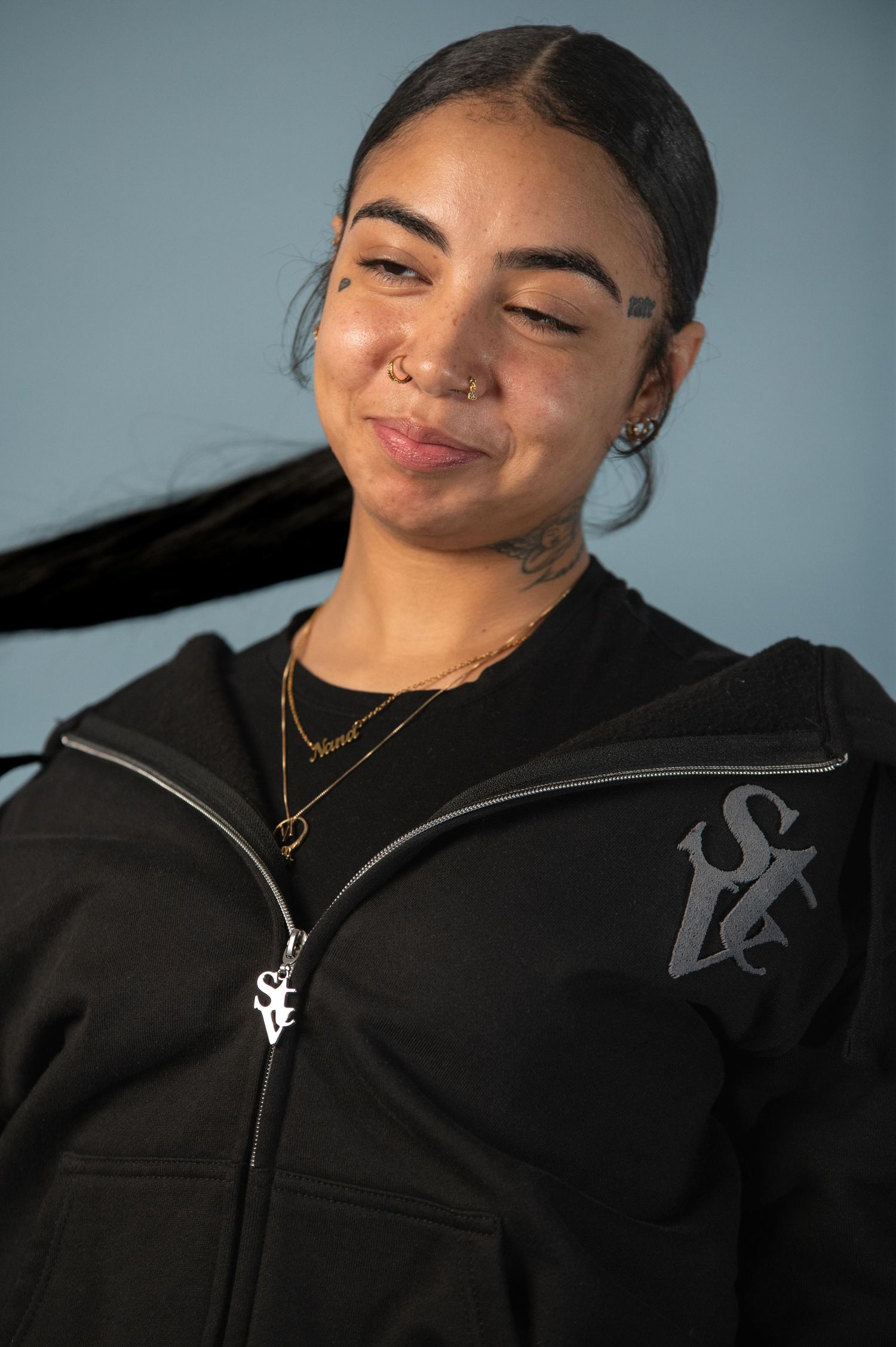Young woman with face tattoos smiles wearing black zip-up hoodie.