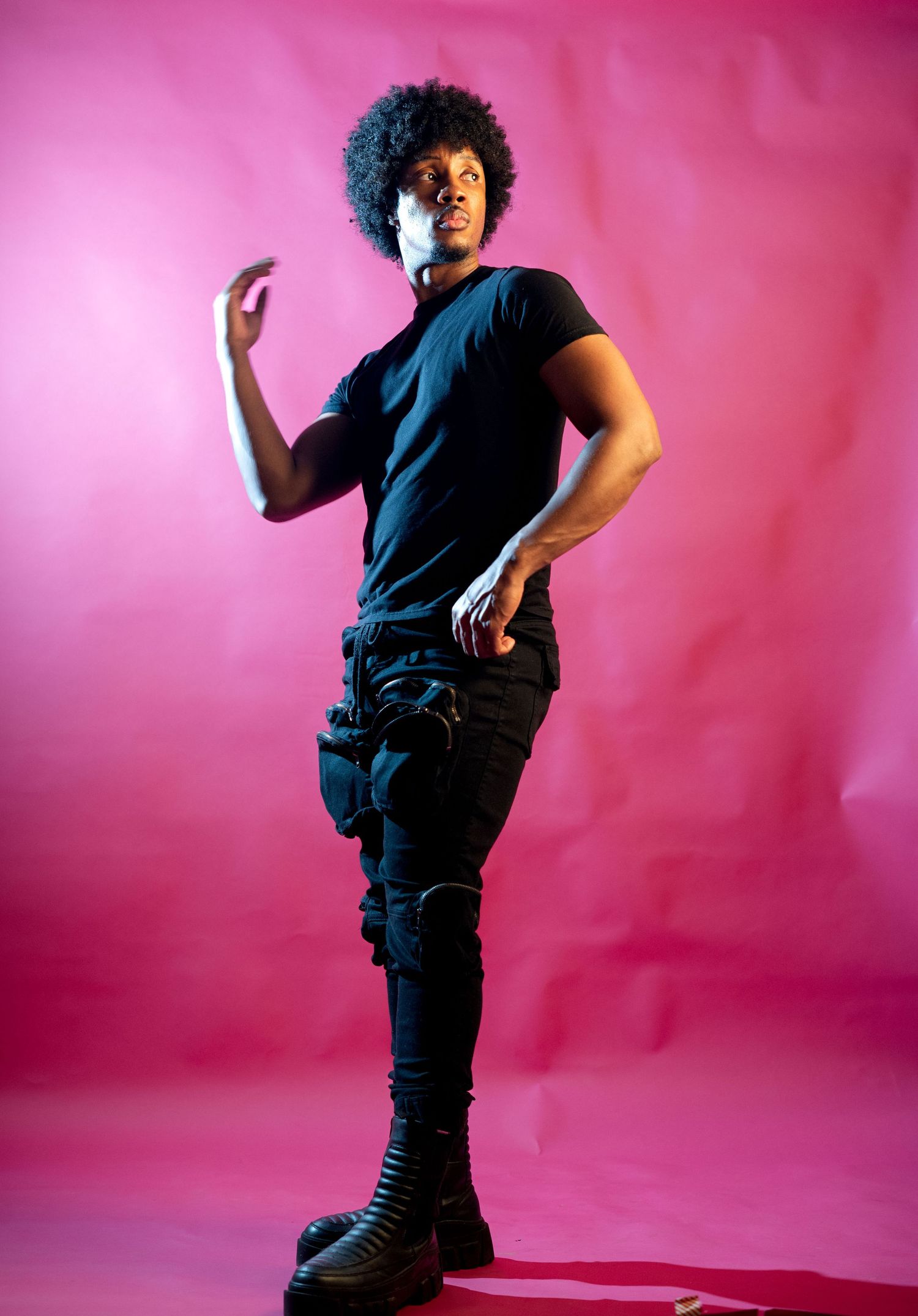 Man in black outfit with combat boots poses confidently against vibrant pink studio backdrop.