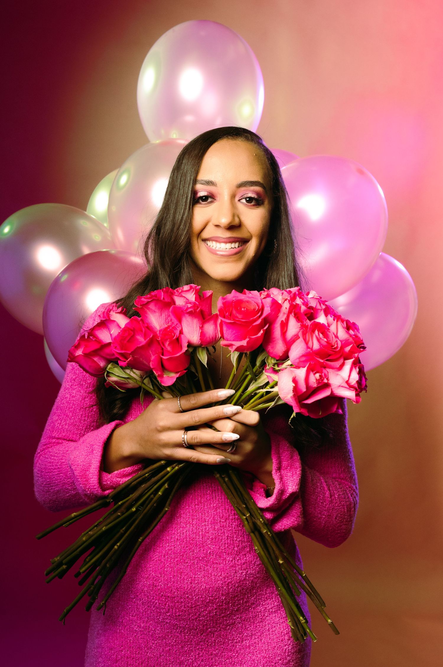 Joyful woman in pink tweed jacket embraces large bouquet of pink roses with balloons behind her.