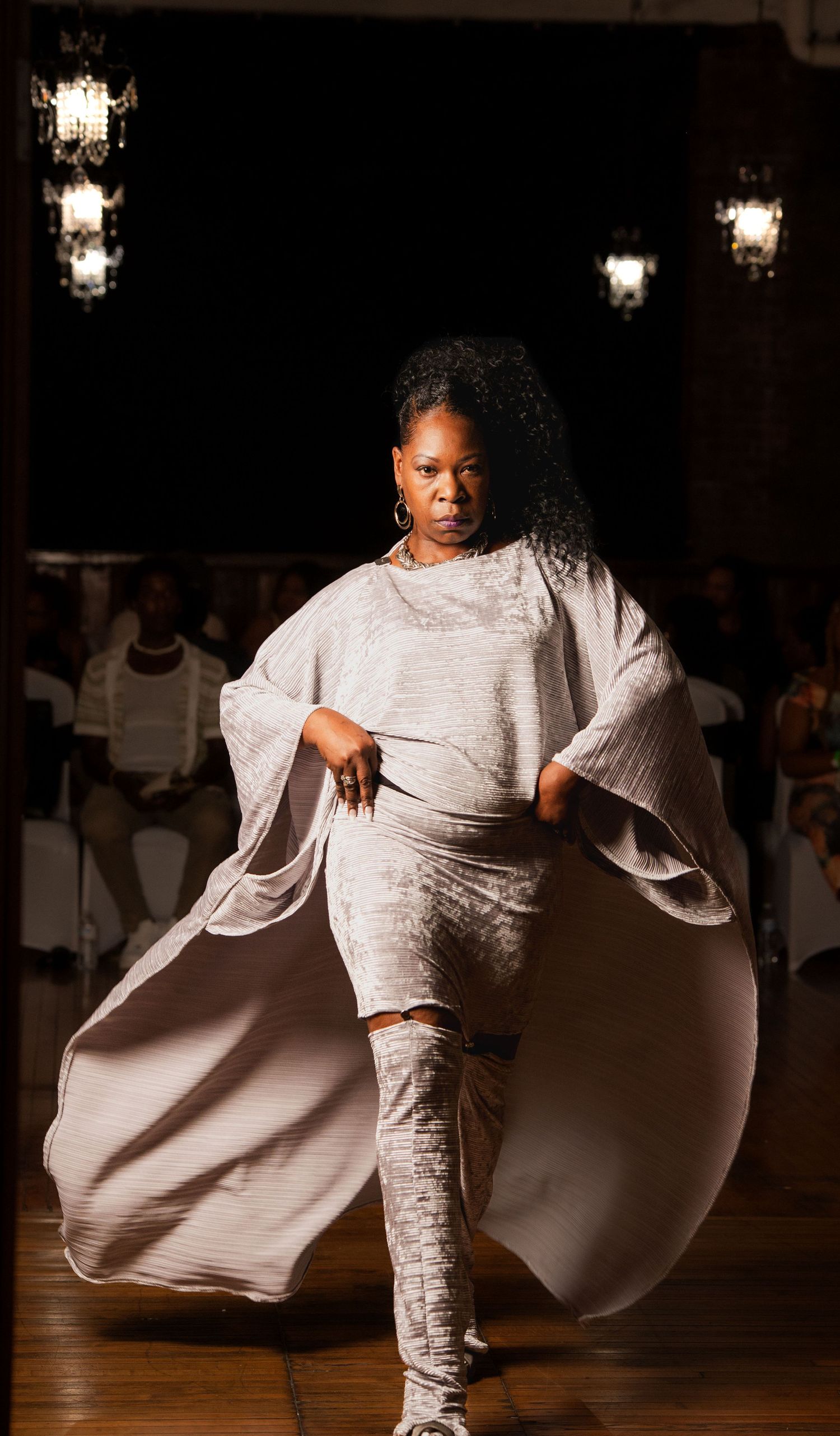 Plus-size model walks runway in silver-gray flowing cape ensemble at fashion show.