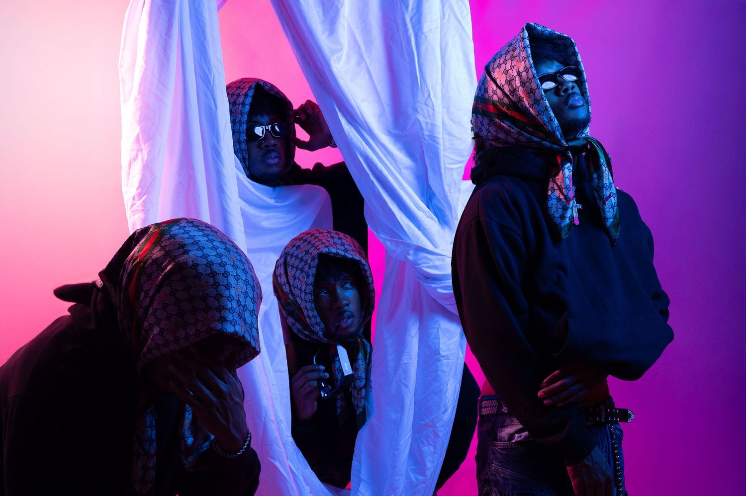 Three masked figures in dark clothing pose dramatically under purple and pink neon lighting.