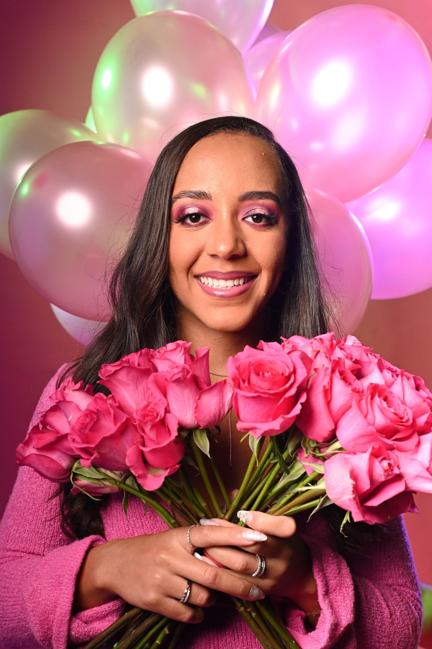 Smiling woman in pink sweater holds bouquet of pink roses with balloons in background.