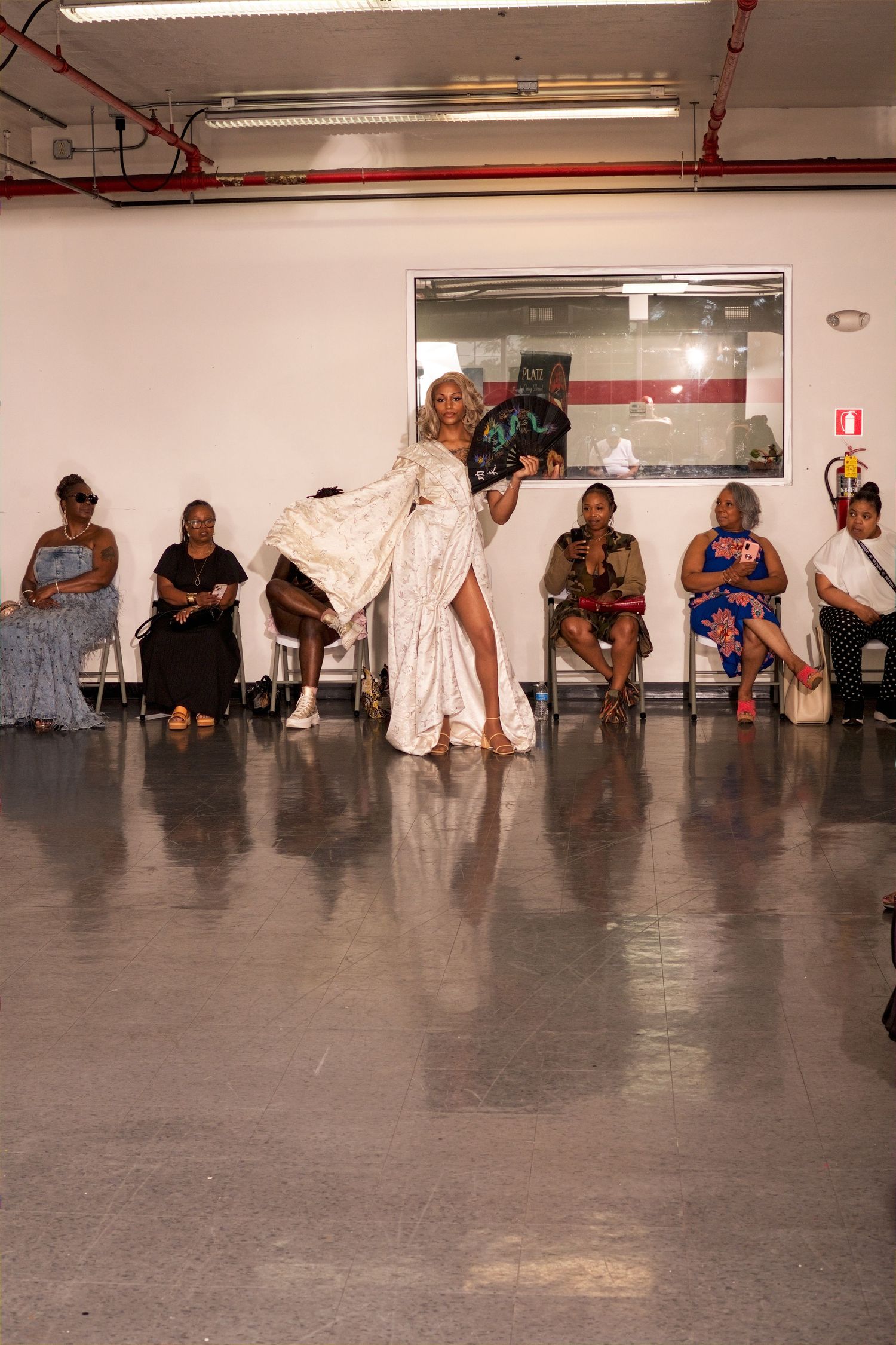 Female model in elegant white cape gown walks runway before seated audience at fashion show.
