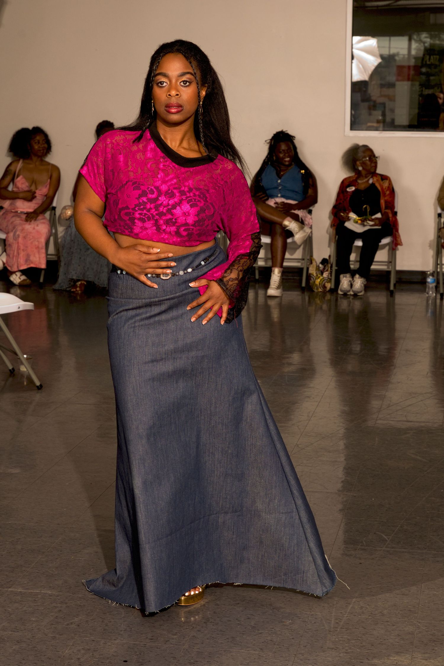 Female model in pink lace top and long denim skirt poses on fashion show runway.