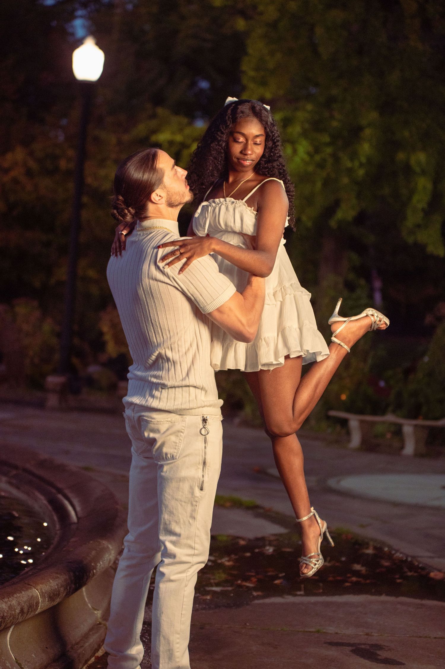 Man lifts woman in white mini dress near a fountain in a romantic evening park setting.
