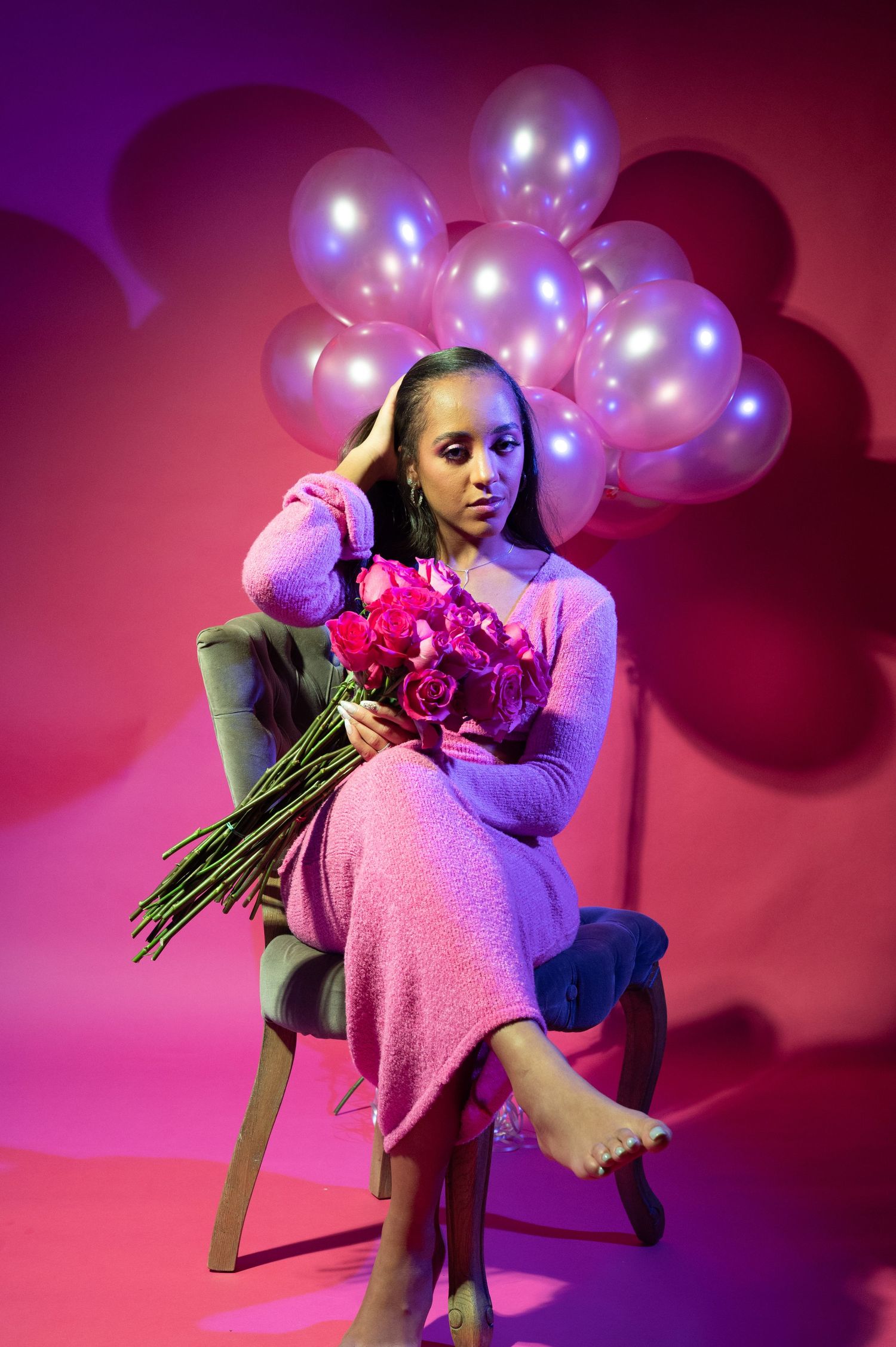 Woman in pink dress sitting on chair holding bouquet of pink roses with balloons in background.
