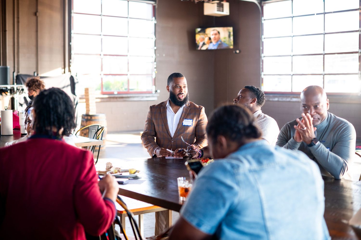 Well-dressed man in brown suit leads a discussion with a group at a restaurant table.