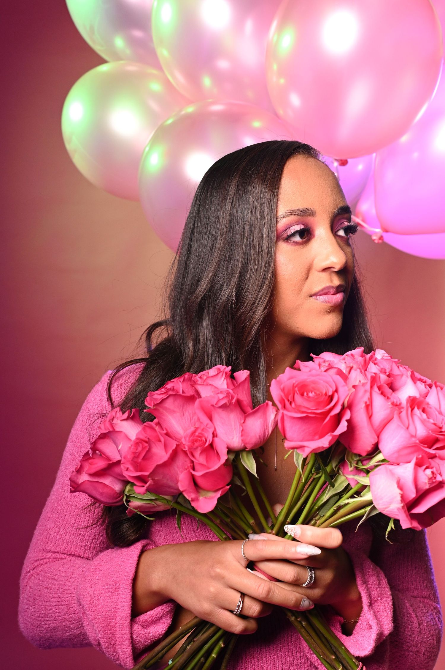 Woman in pink sweater holds bouquet of pink roses with glowing balloons in background.