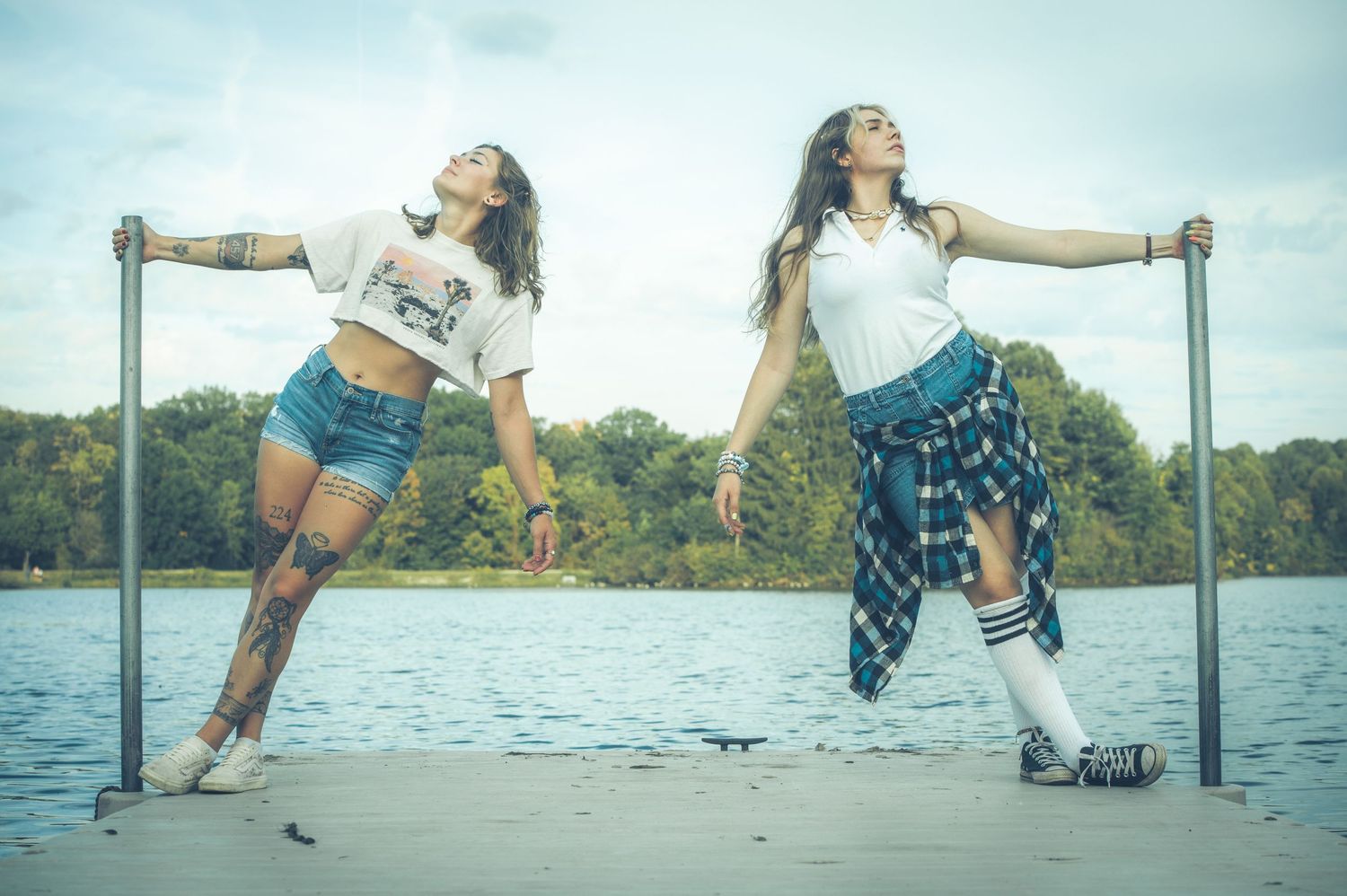 Two women pose dynamically on a lakeside dock, wearing casual white tees and denim outfits.