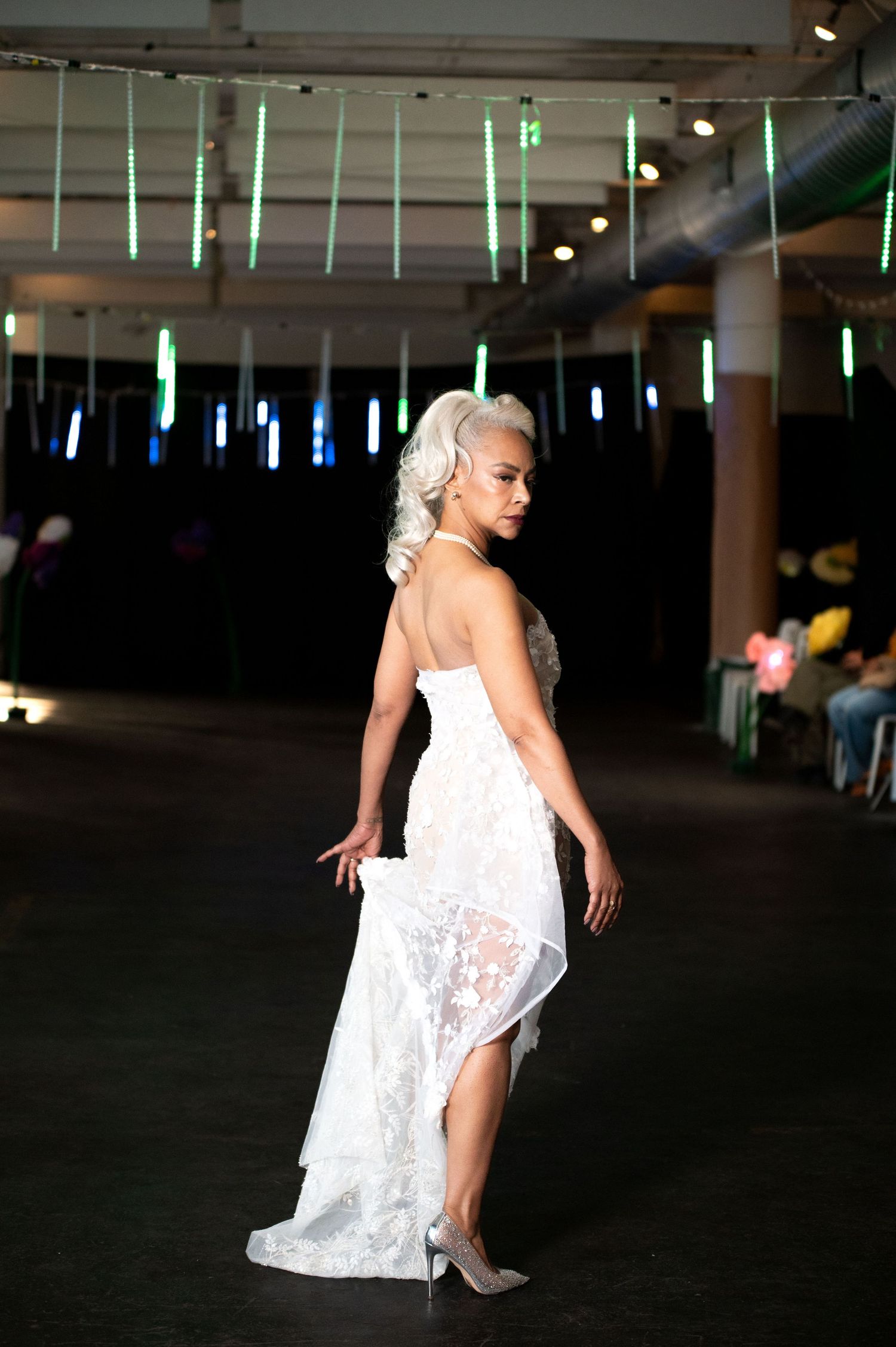 Woman in white lace strapless gown with silver heels walks runway under green neon lights.