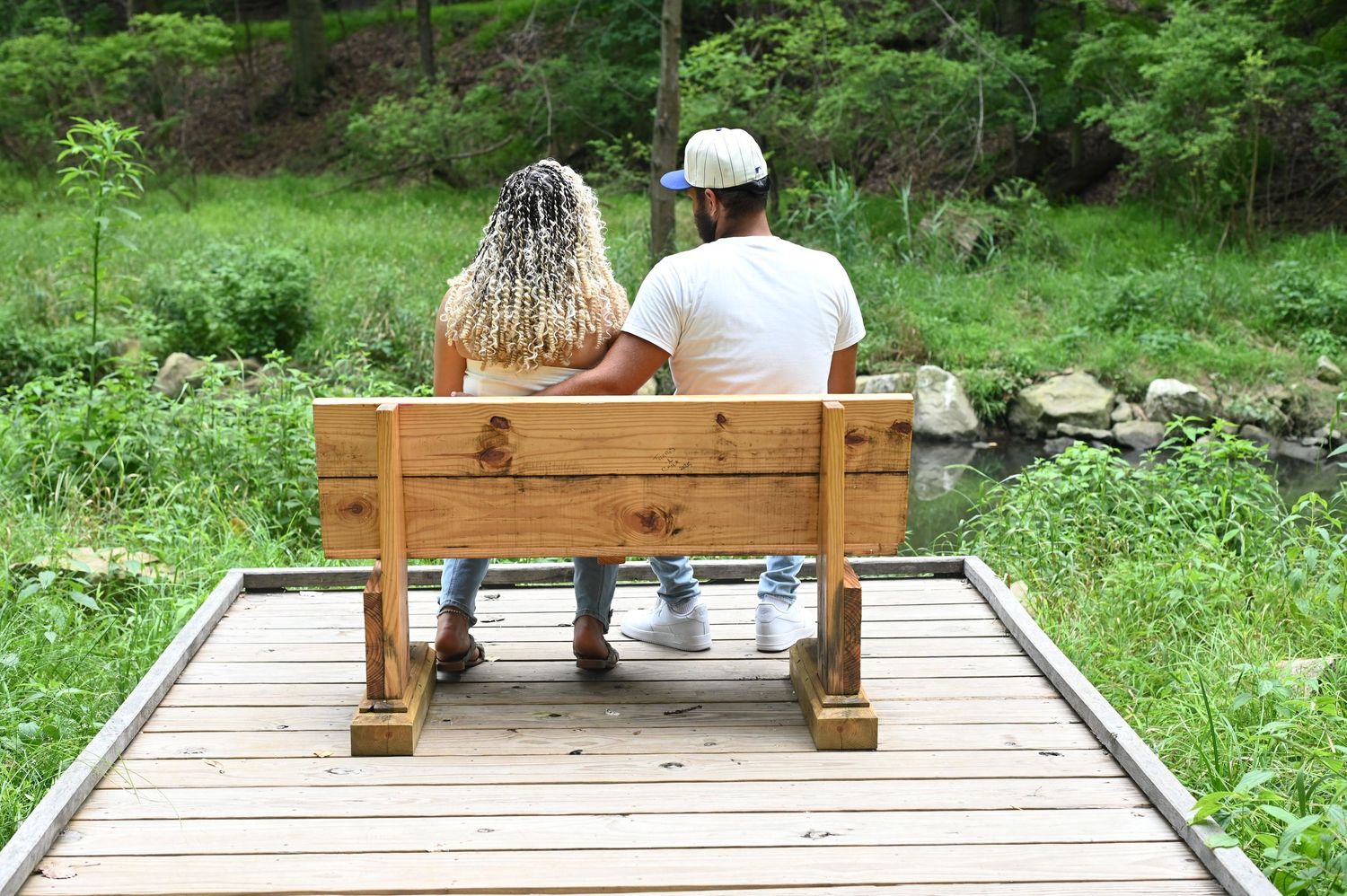 Couple sitting on wooden bench viewed from behind, surrounded by lush green forest landscape.