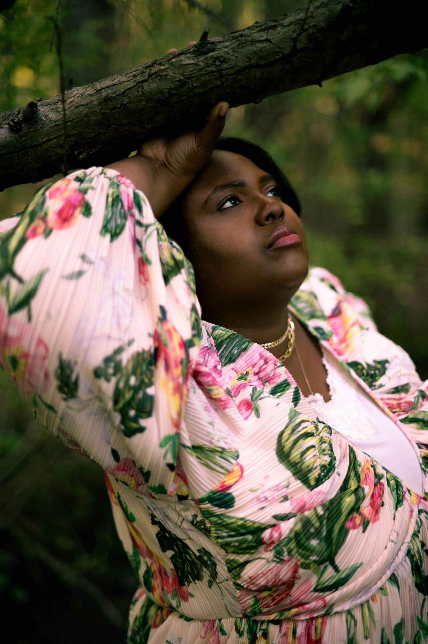 Woman in floral pink dress leans against tree branch outdoors.