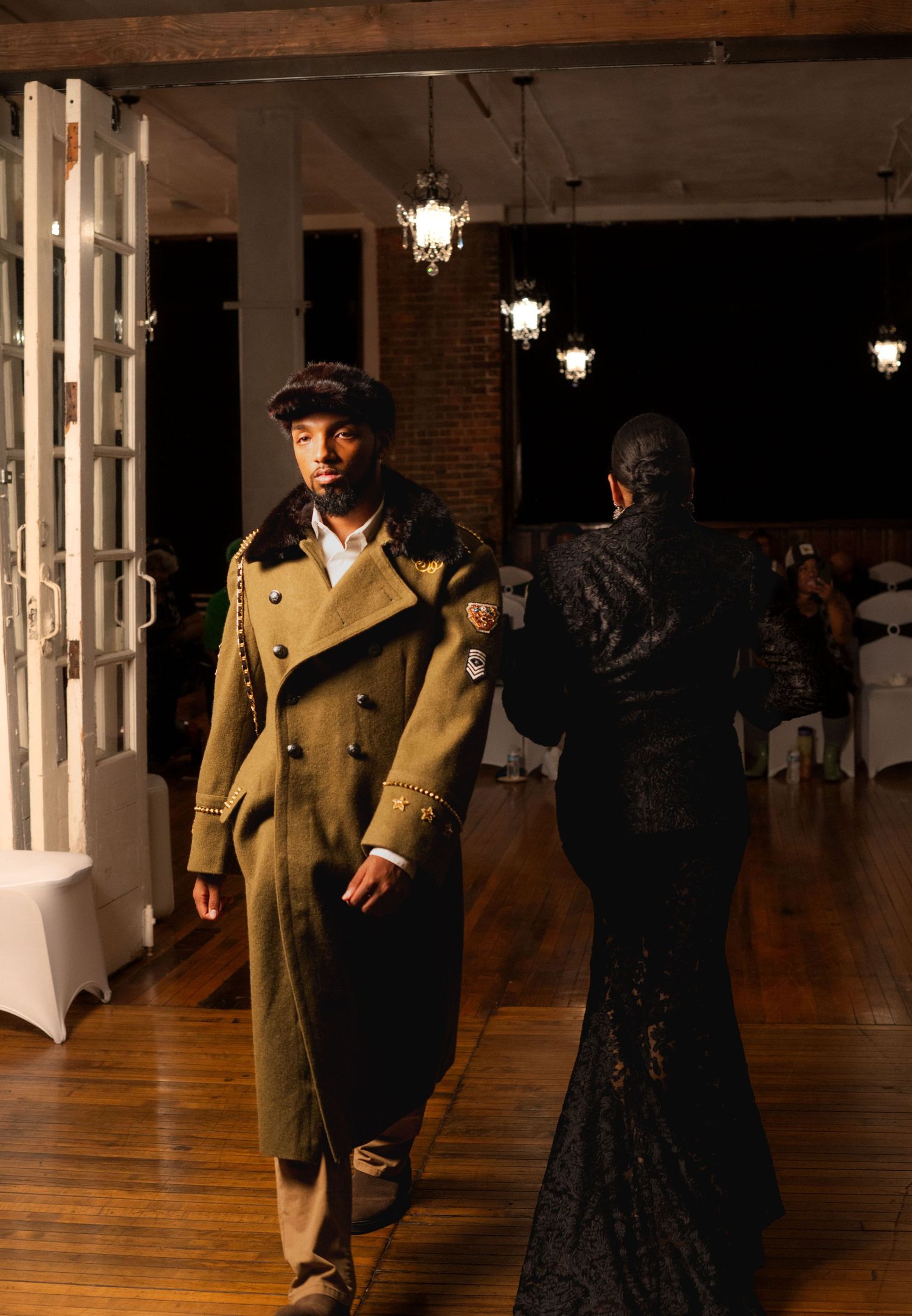 Male model in olive military coat with fur collar walks runway at fashion show.