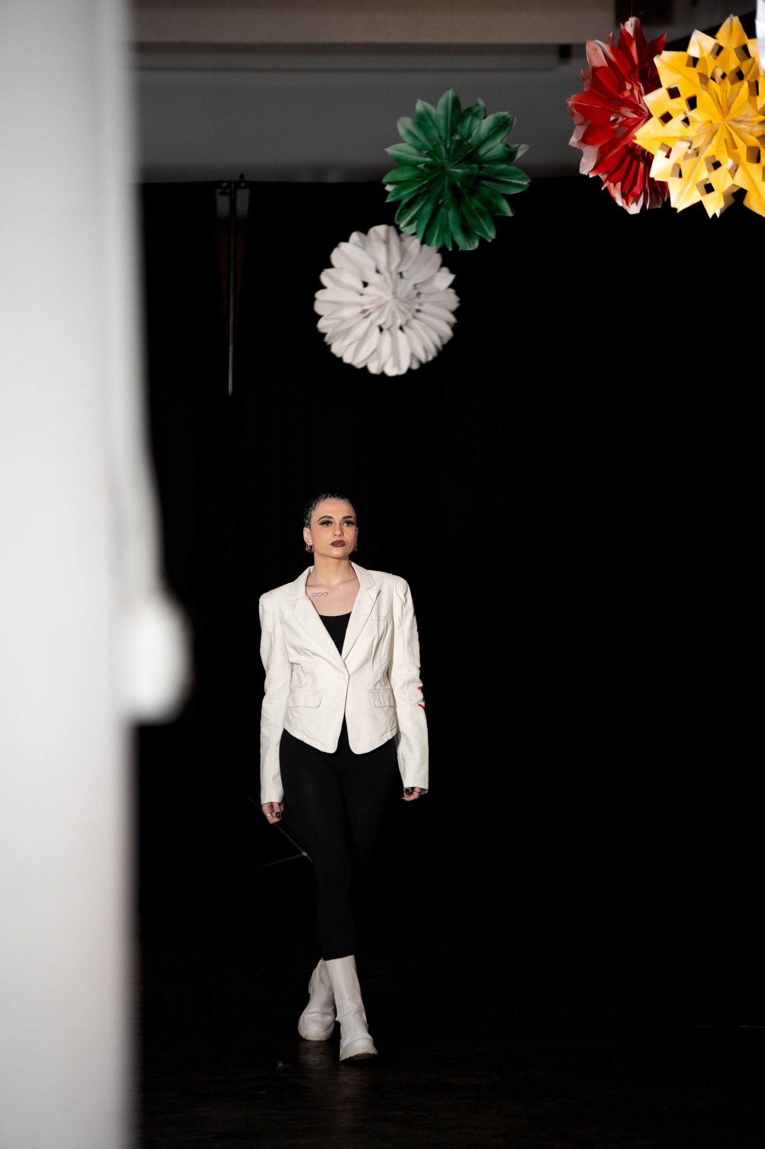 Female model walks runway in white blazer and black pants under decorative paper flowers.