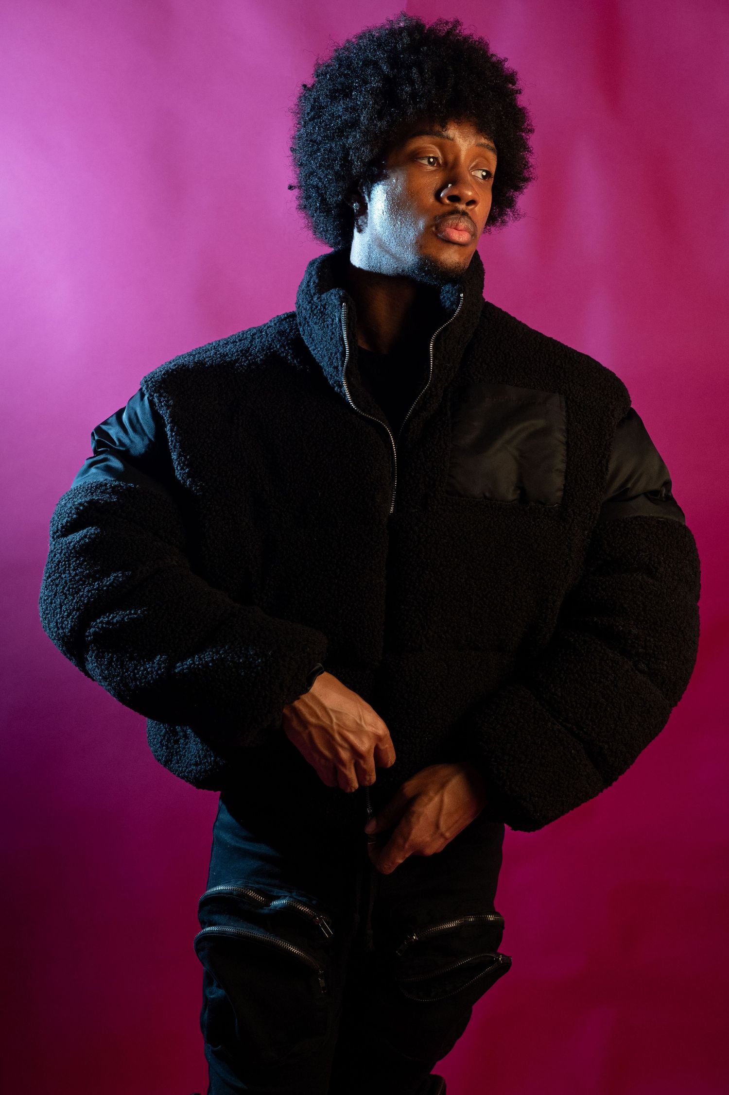 Man with afro in black sherpa jacket poses on pink background.