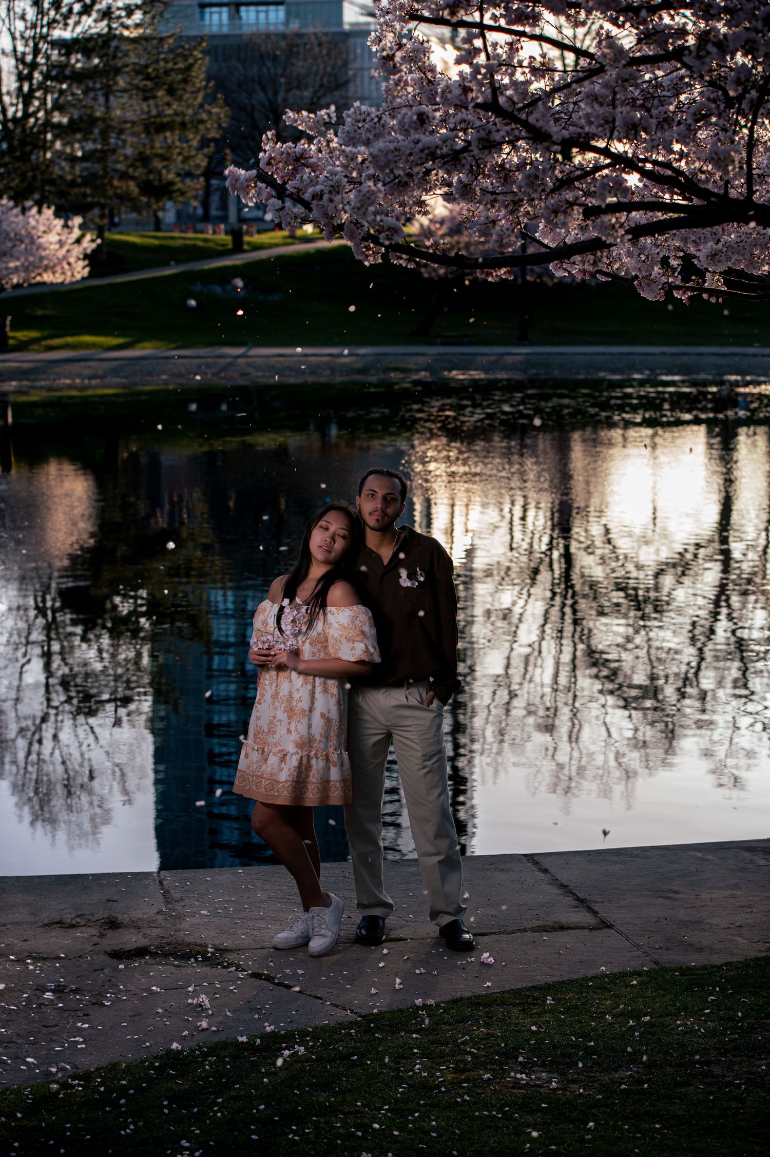Couple embraces romantically by a reflective pond under blooming cherry blossom trees at dusk.