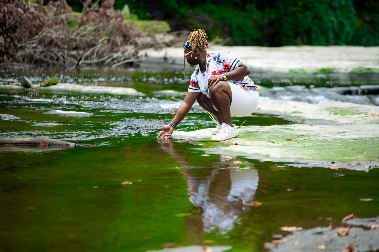 Young woman in white outfit crouches by a green algae-covered rocky river in nature.