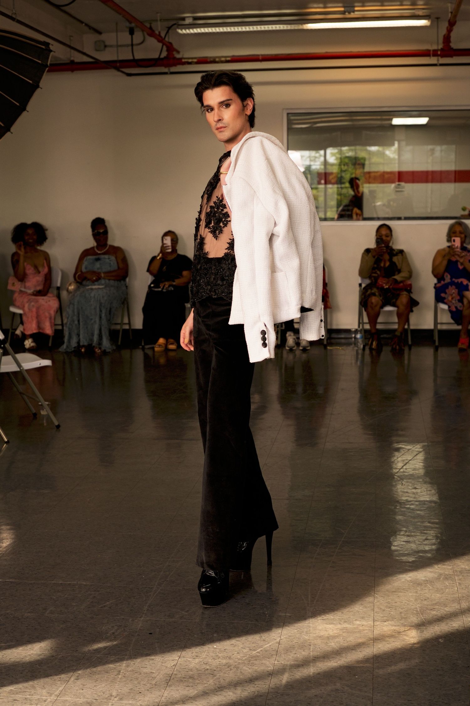 Model walks runway in white blazer, black lace bodysuit, and wide-leg trousers with platform heels at fashion show.