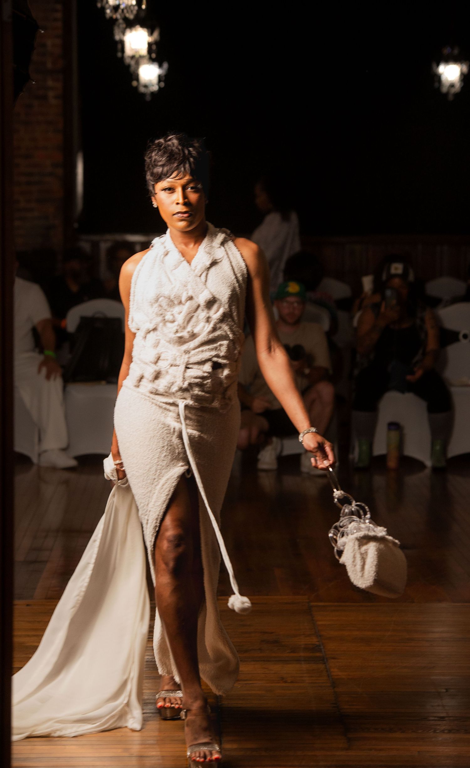 Model walks runway in white textured halter gown with fringe details at fashion show.