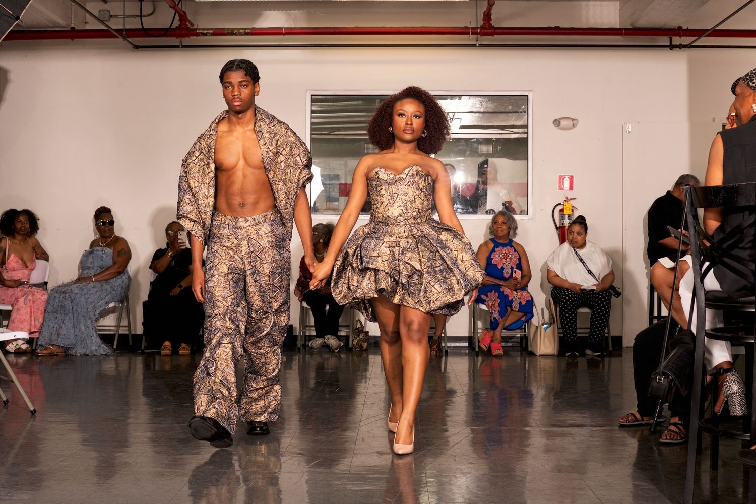Two models walk runway together in matching metallic printed outfits at indoor fashion show.