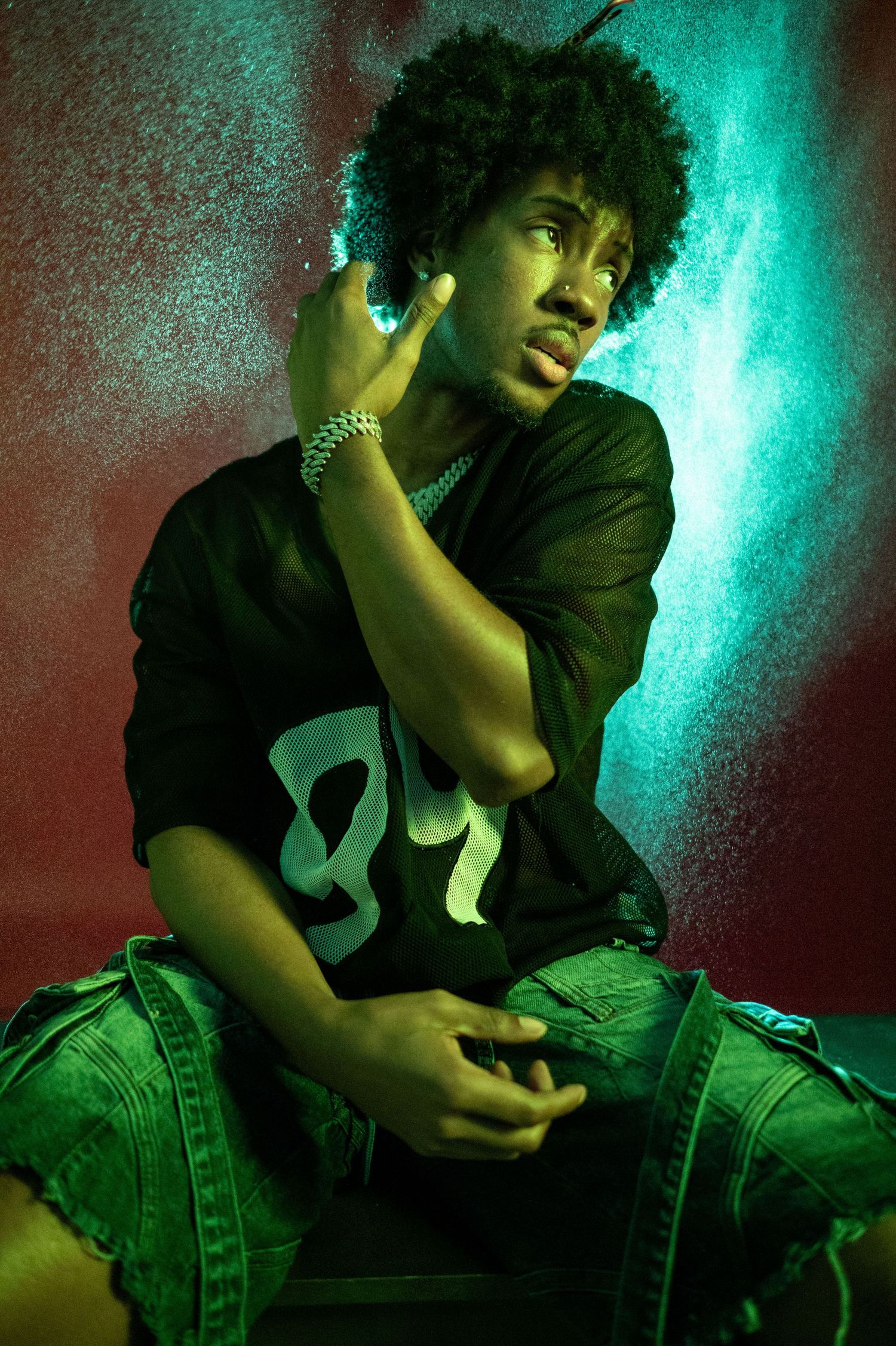 Young man in black graphic tee and denim shorts poses under dramatic green and red lighting.