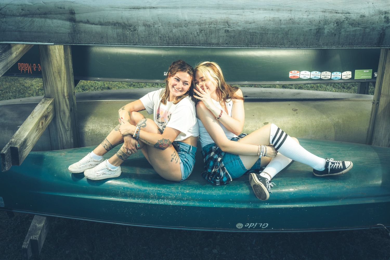 Two young women sit together smiling on a teal bench in casual streetwear outfits.