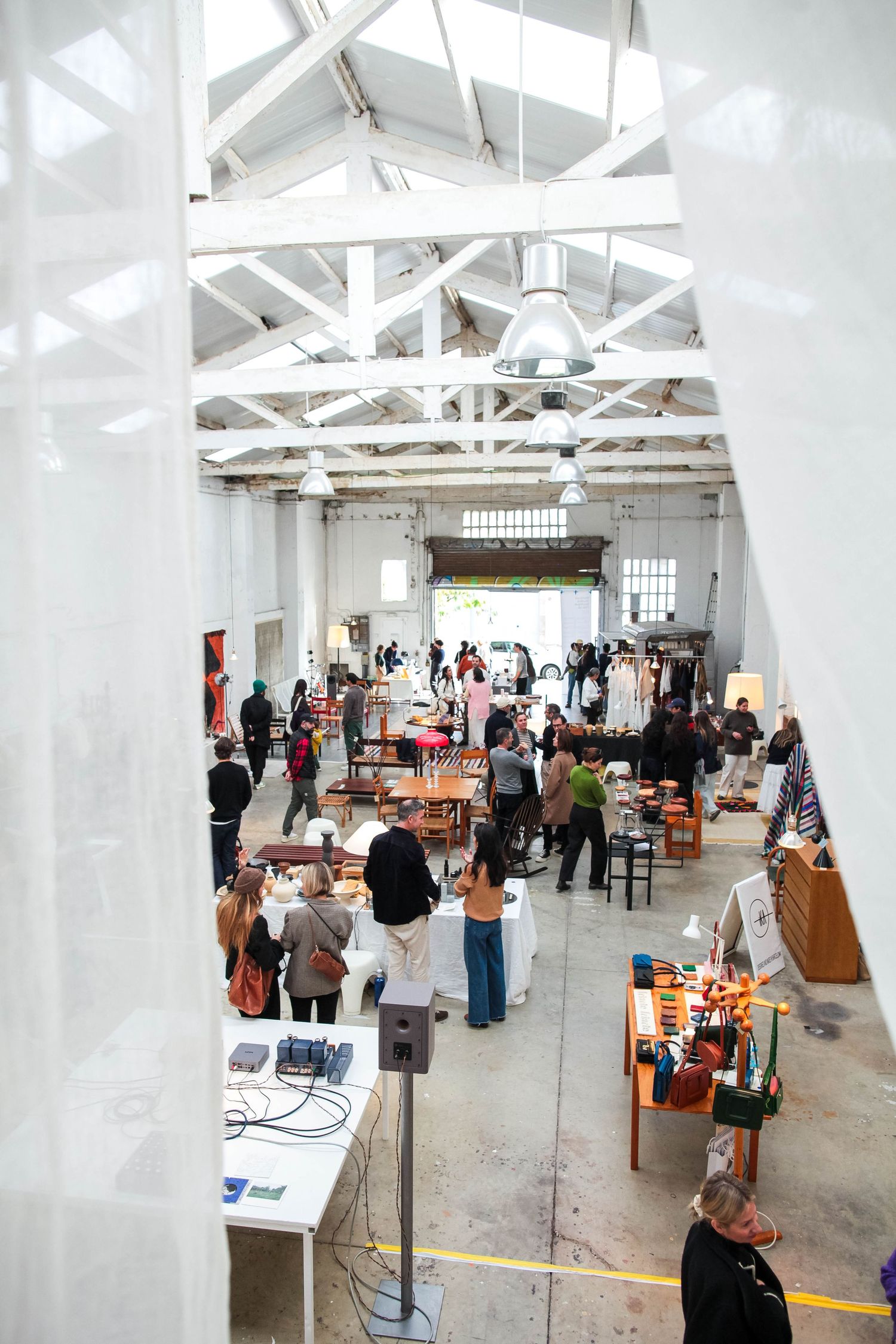 Indoor market or craft fair with white ceiling beams and vendors displaying items on tables.